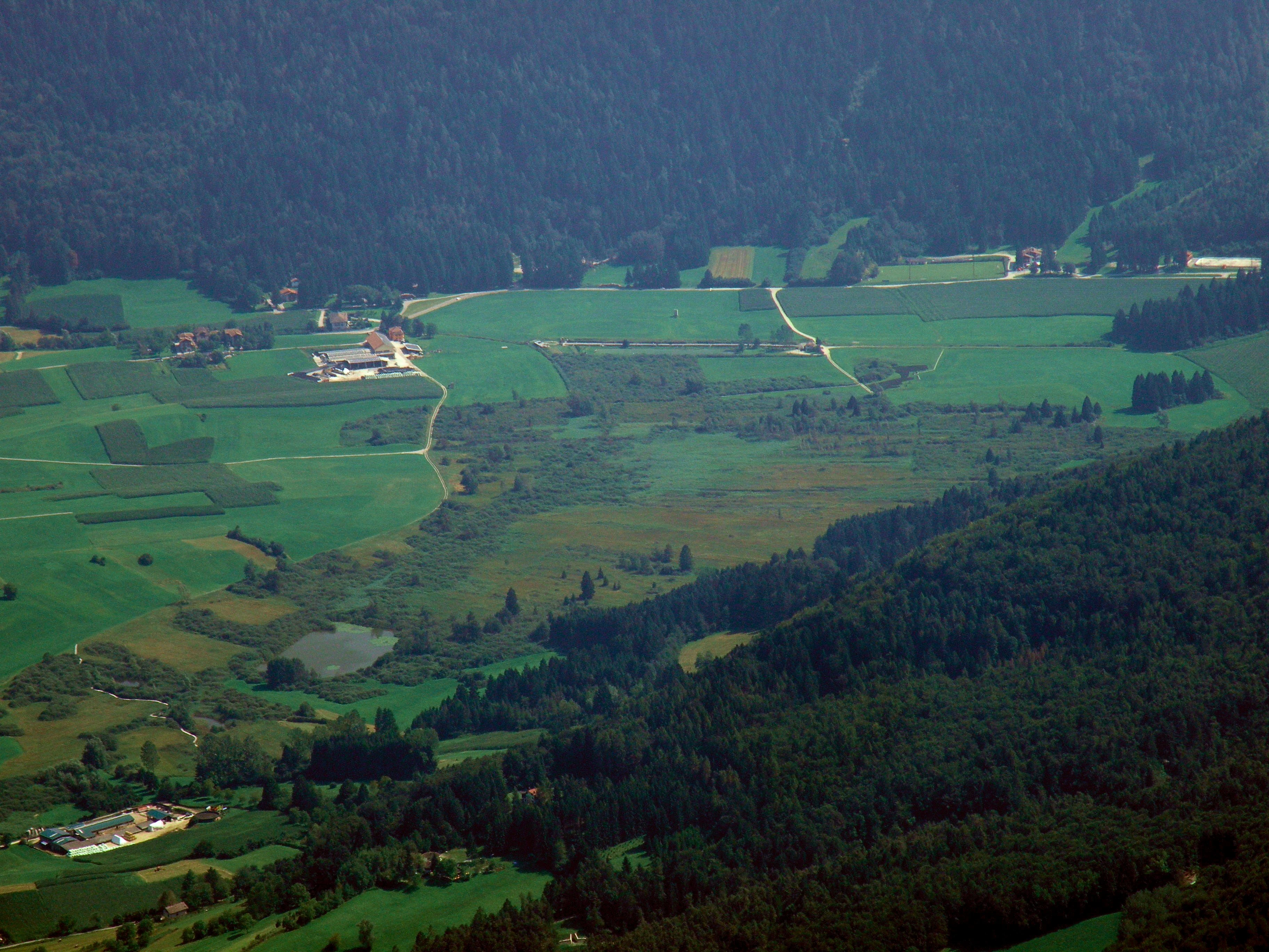 An elevated landscape view of a green valley featuring wetlands and agricultural fields surrounded by forested mountain slopes.