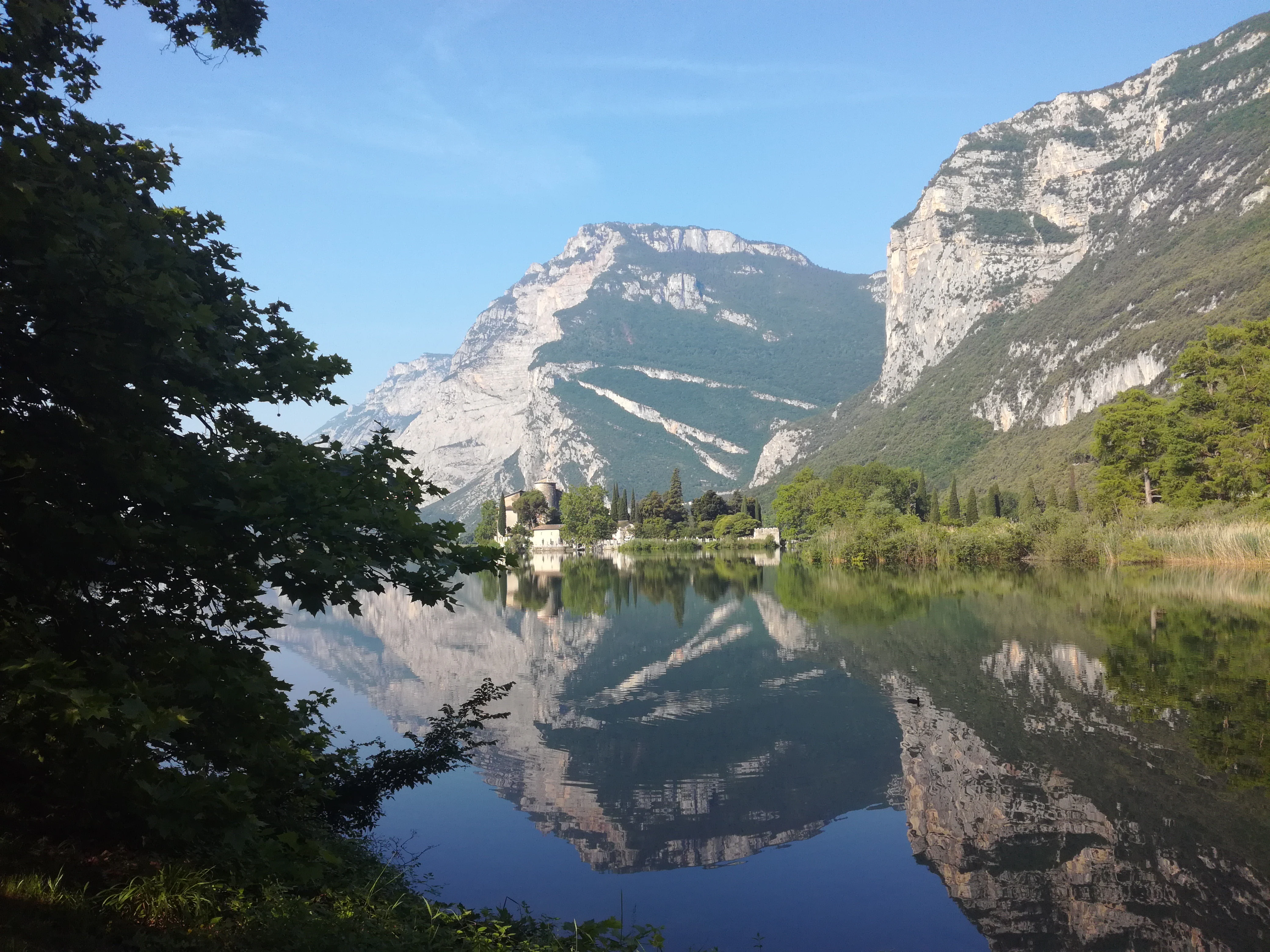 Reflejo de un lago tranquilo que muestra imponentes acantilados de piedra caliza, un pequeño castillo en la distancia y ramas de árboles en primer plano en un día despejado en el Biotopo Lago di Toblino.