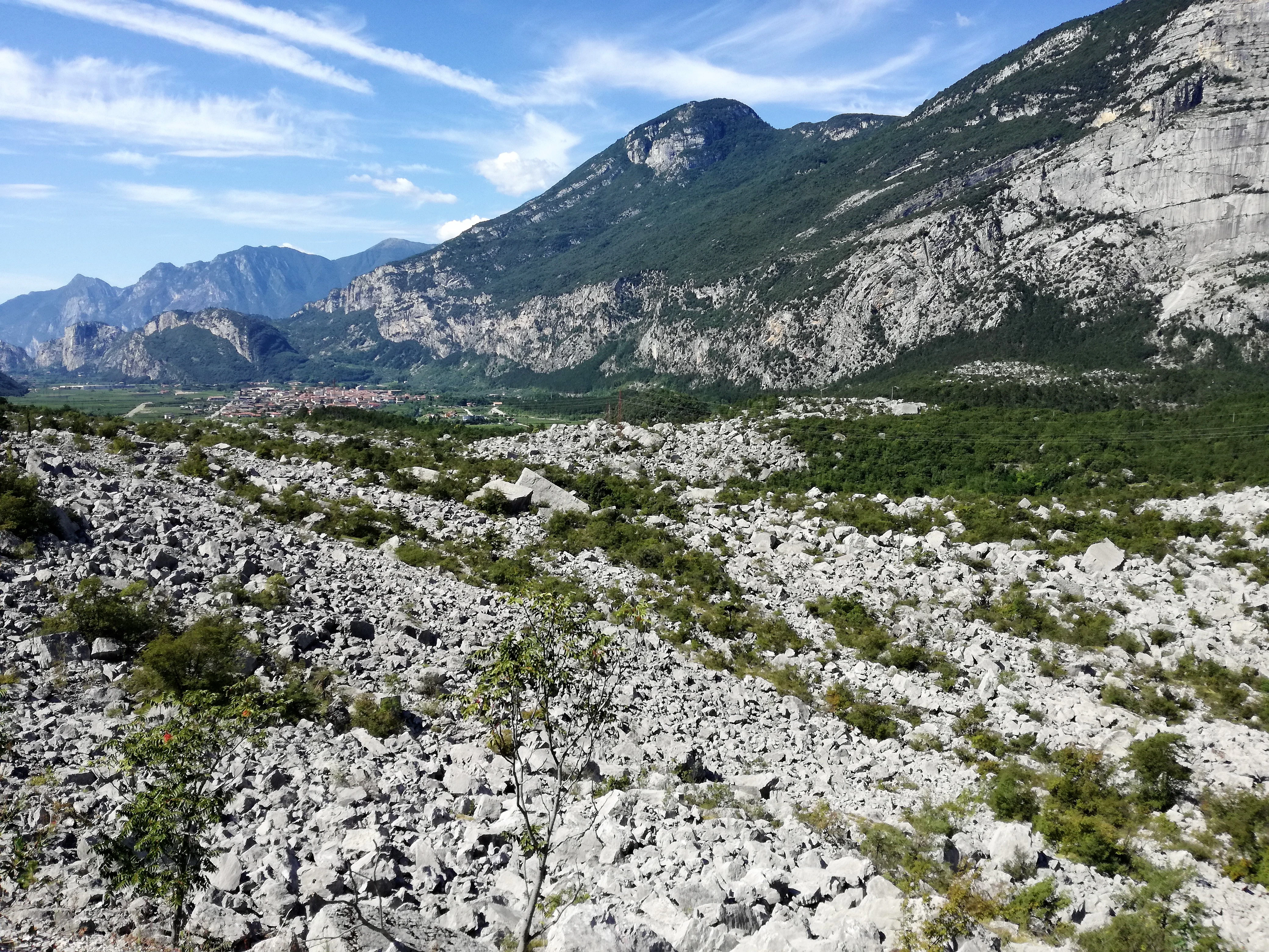 A wide, rocky landscape filled with vast amounts of grey boulders and sparse vegetation, set against a backdrop of steep mountain slopes under a blue sky.