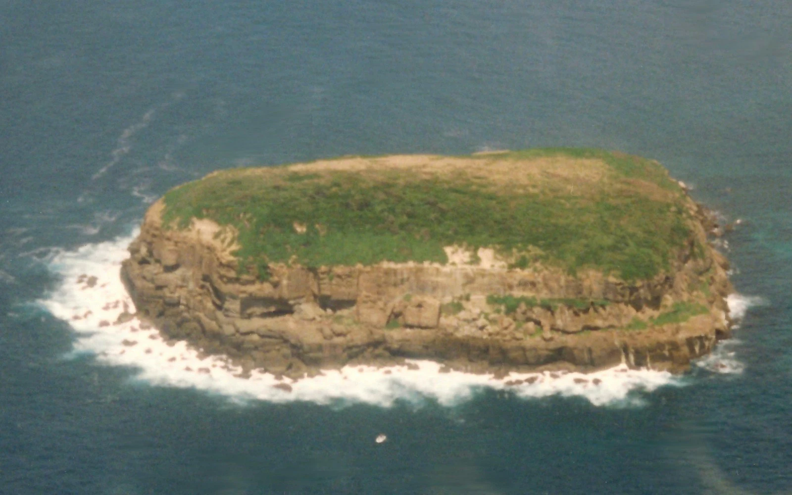 An aerial view of a small, rocky island covered in green vegetation, surrounded by blue ocean water with white surf breaking against its shoreline.