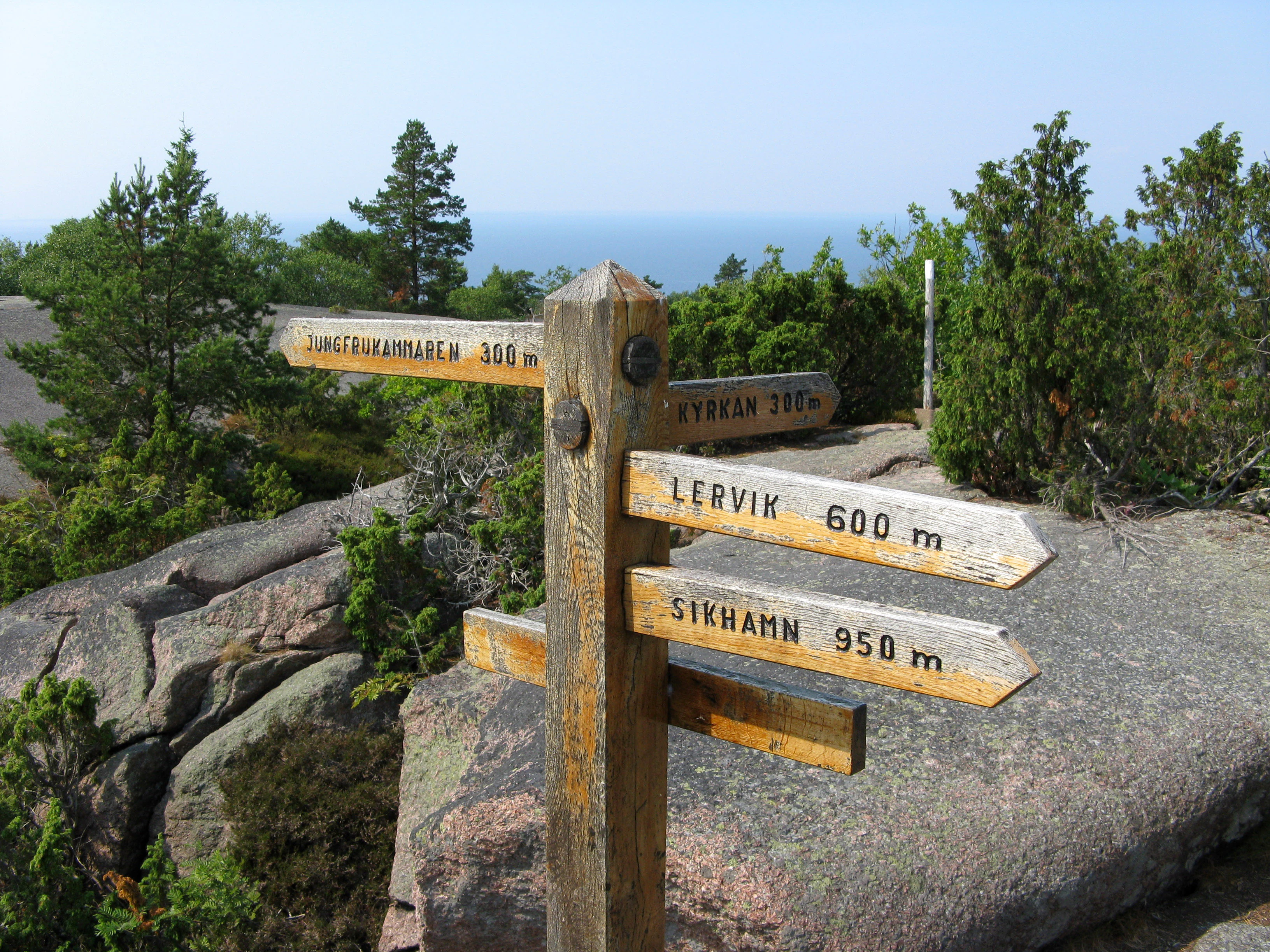 Wooden signpost with multiple directional signs displaying distances to Lervik (600 m), Sirkhamn (950 m), and other locations, set against rocky terrain with trees and distant sea view