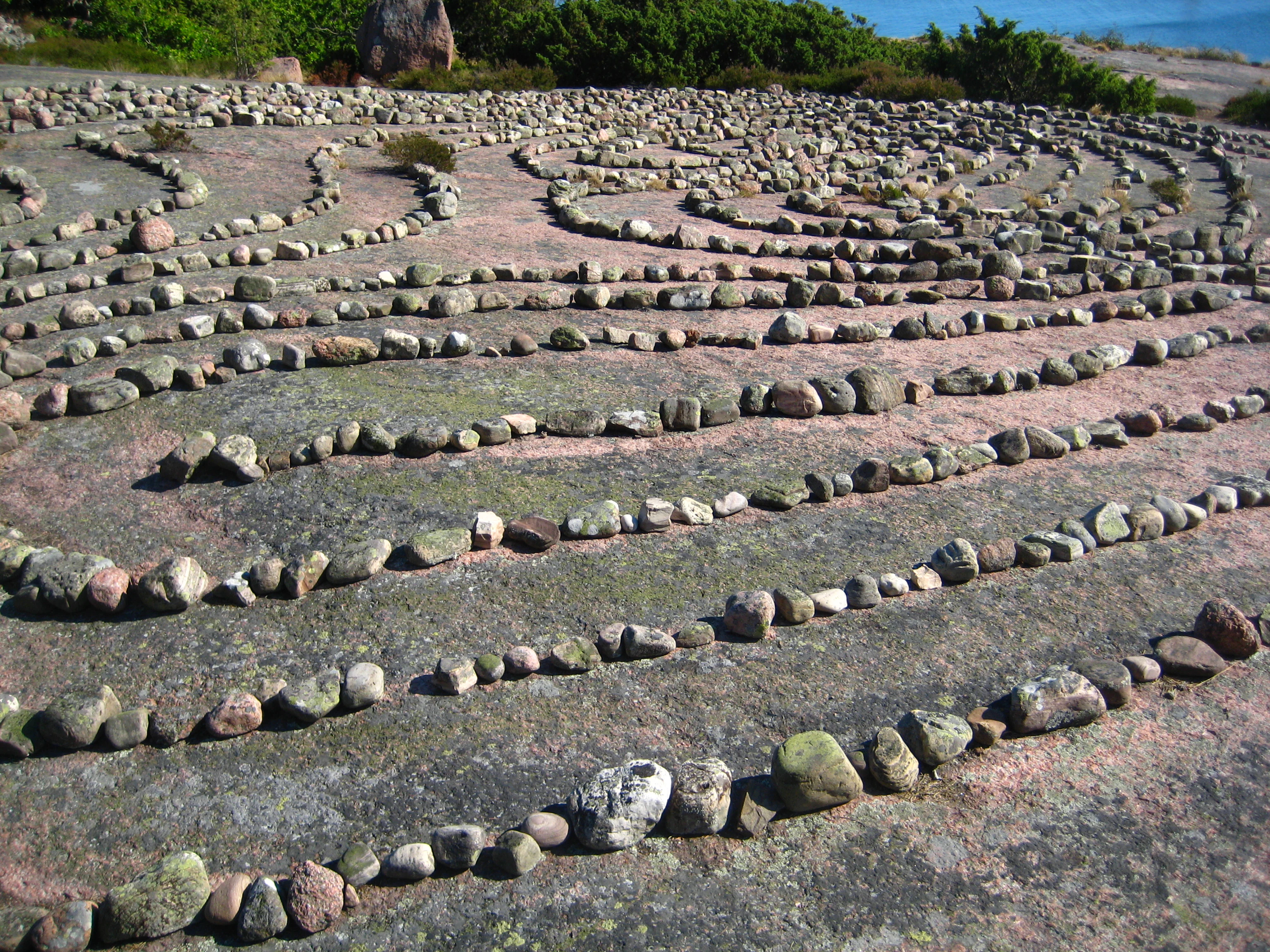 A stone labyrinth composed of arranged rocks on a large flat rock surface with patches of green moss, surrounded by green vegetation in the background.