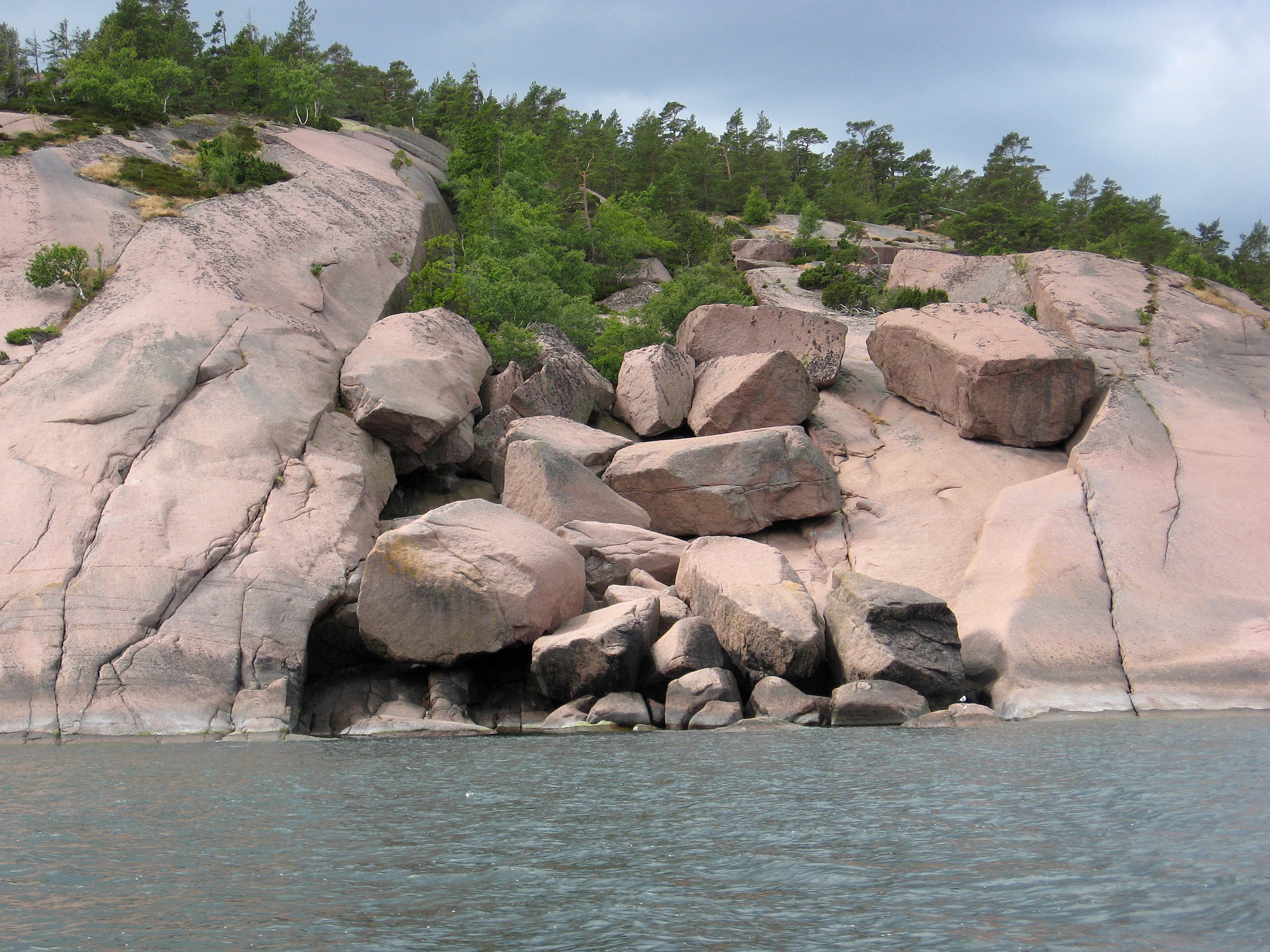 Rocky cape with large boulders and calm water in the foreground, surrounded by forested cliffs under a cloudy sky.