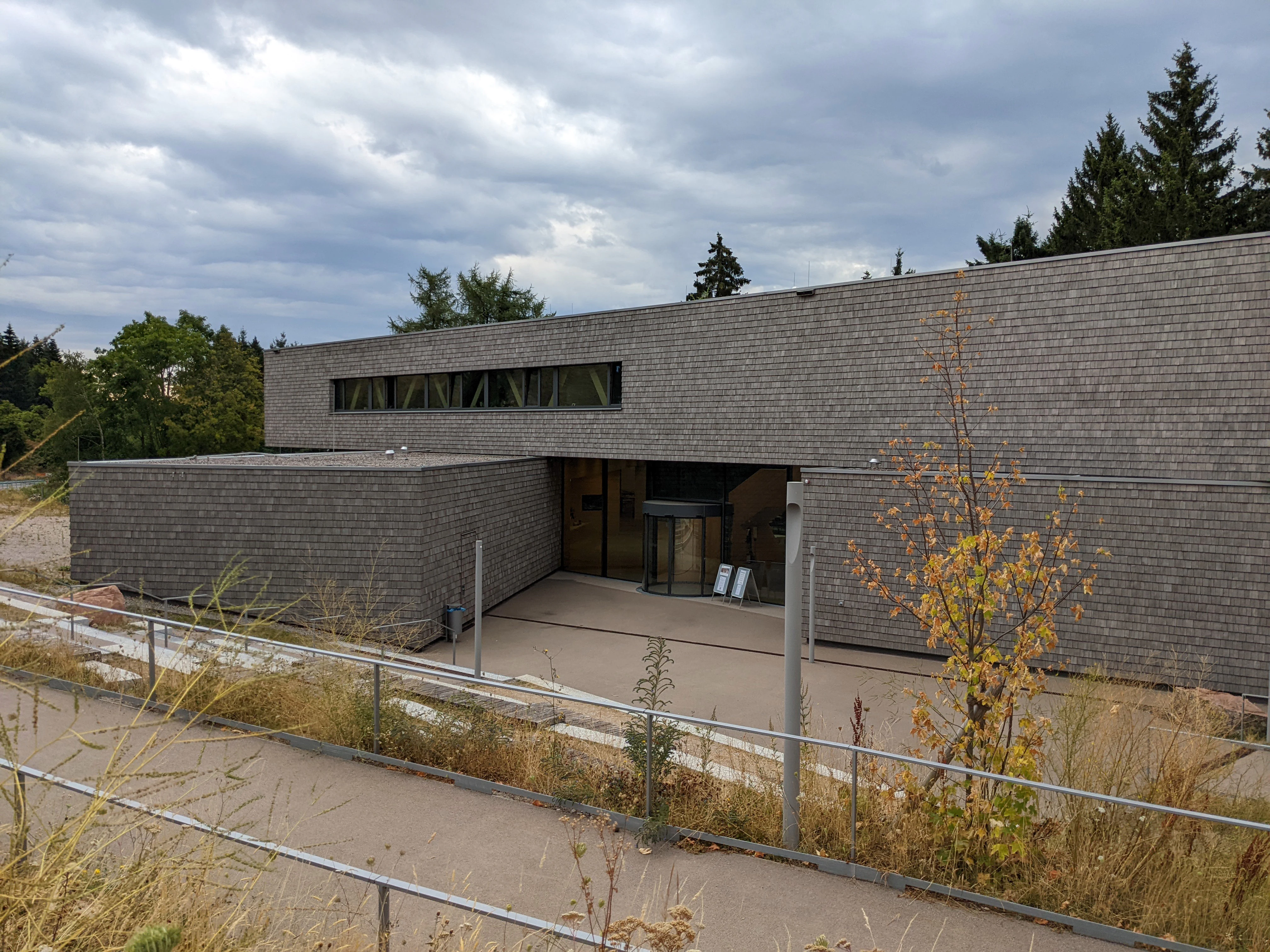 Gray brick building with large windows and entrance area, metal fence, vegetation, and cloudy sky in background