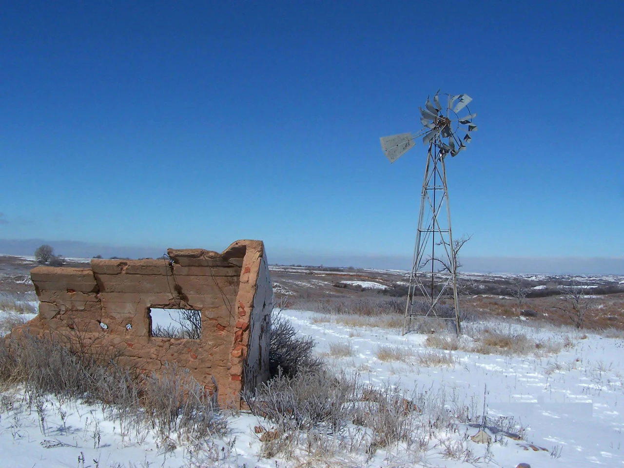 Un molino de viento metálico y desgastado junto a los restos de una pequeña construcción de adobe en ruinas, en una pradera abierta cubierta de nieve bajo un cielo azul despejado.