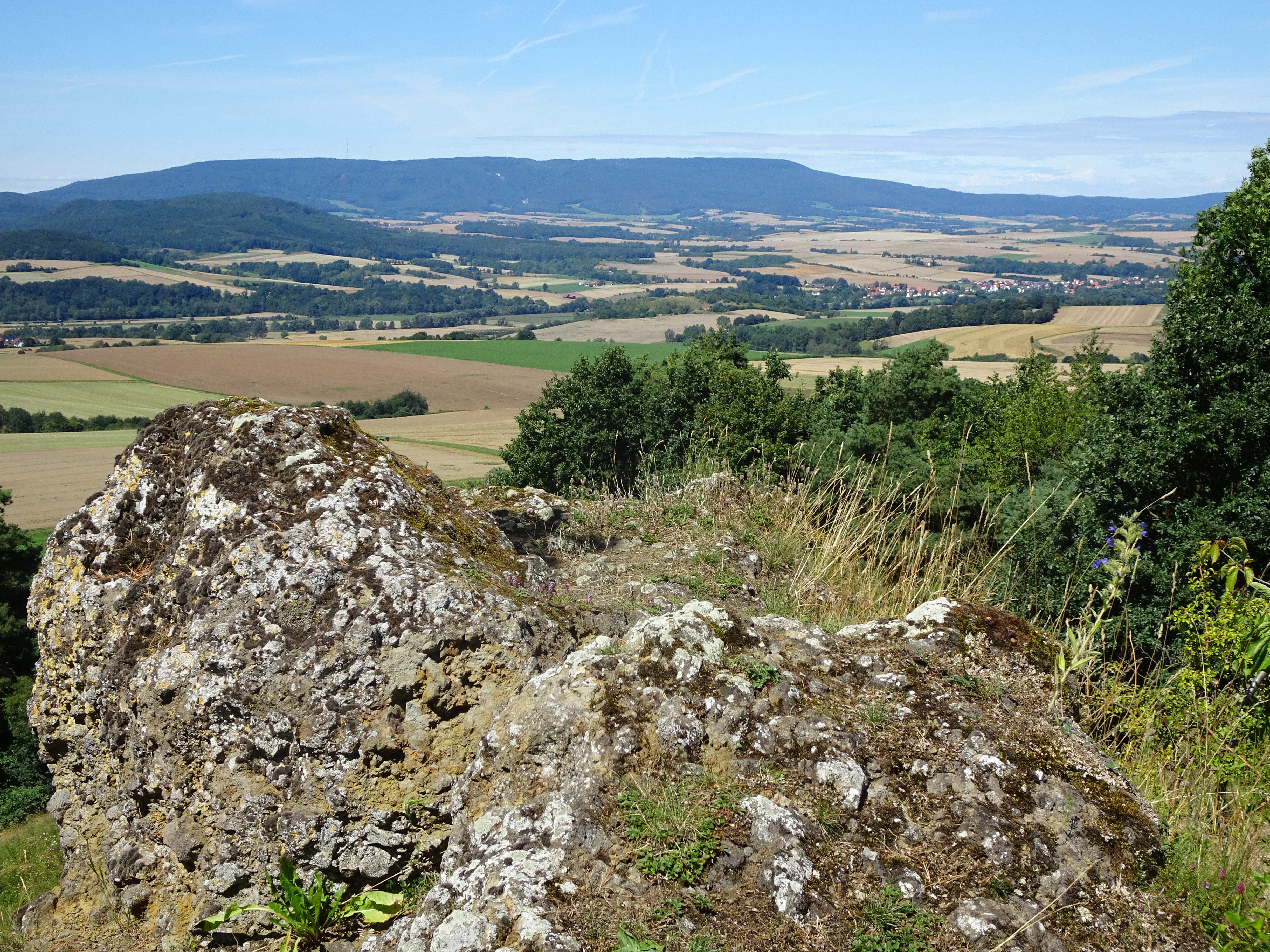 A high angle view over a rolling rural landscape with agricultural fields and distant hills, with moss-covered, textured volcanic basalt boulders in the immediate foreground.