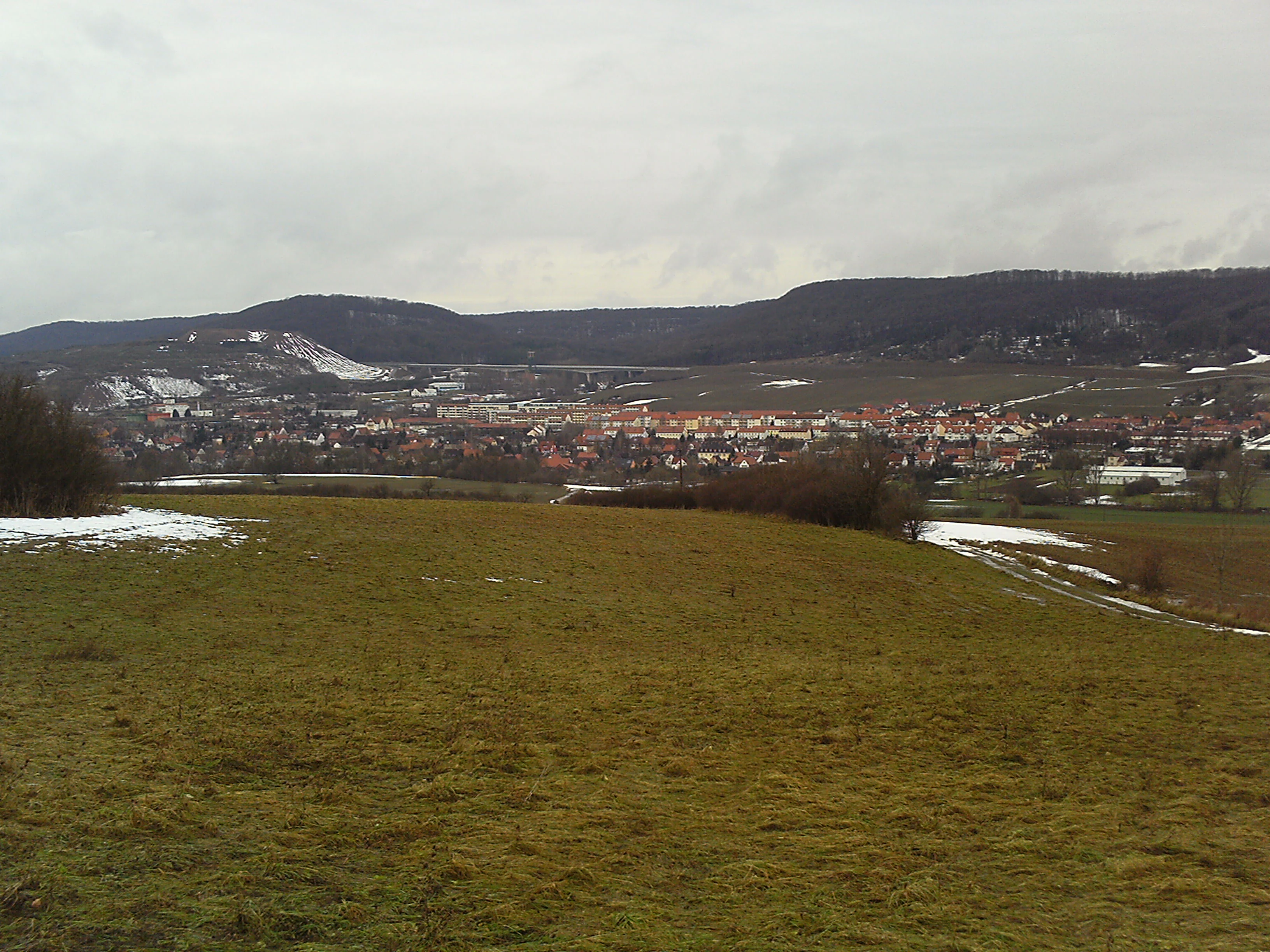 Una vista amplia del paisaje con un prado verde y parcialmente cubierto de nieve en primer plano que conduce al pueblo de Sollstedt y a la línea de la cresta de los Bleicheröder Berge al fondo, bajo un cielo nublado.