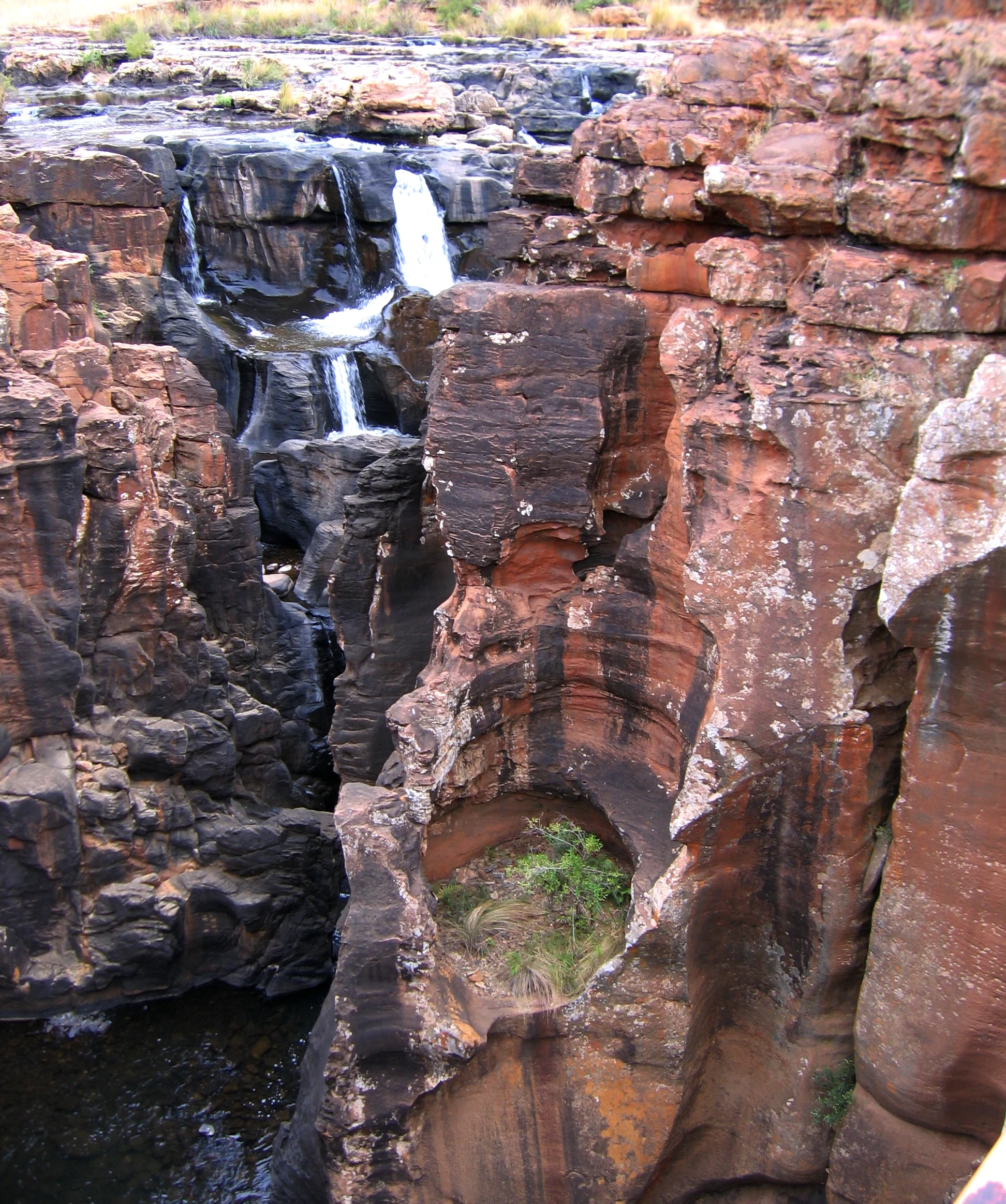 Eroded red and brown sandstone rock formations featuring deep, rounded cylindrical holes carved by water, with a small waterfall flowing into a dark pool in the background.