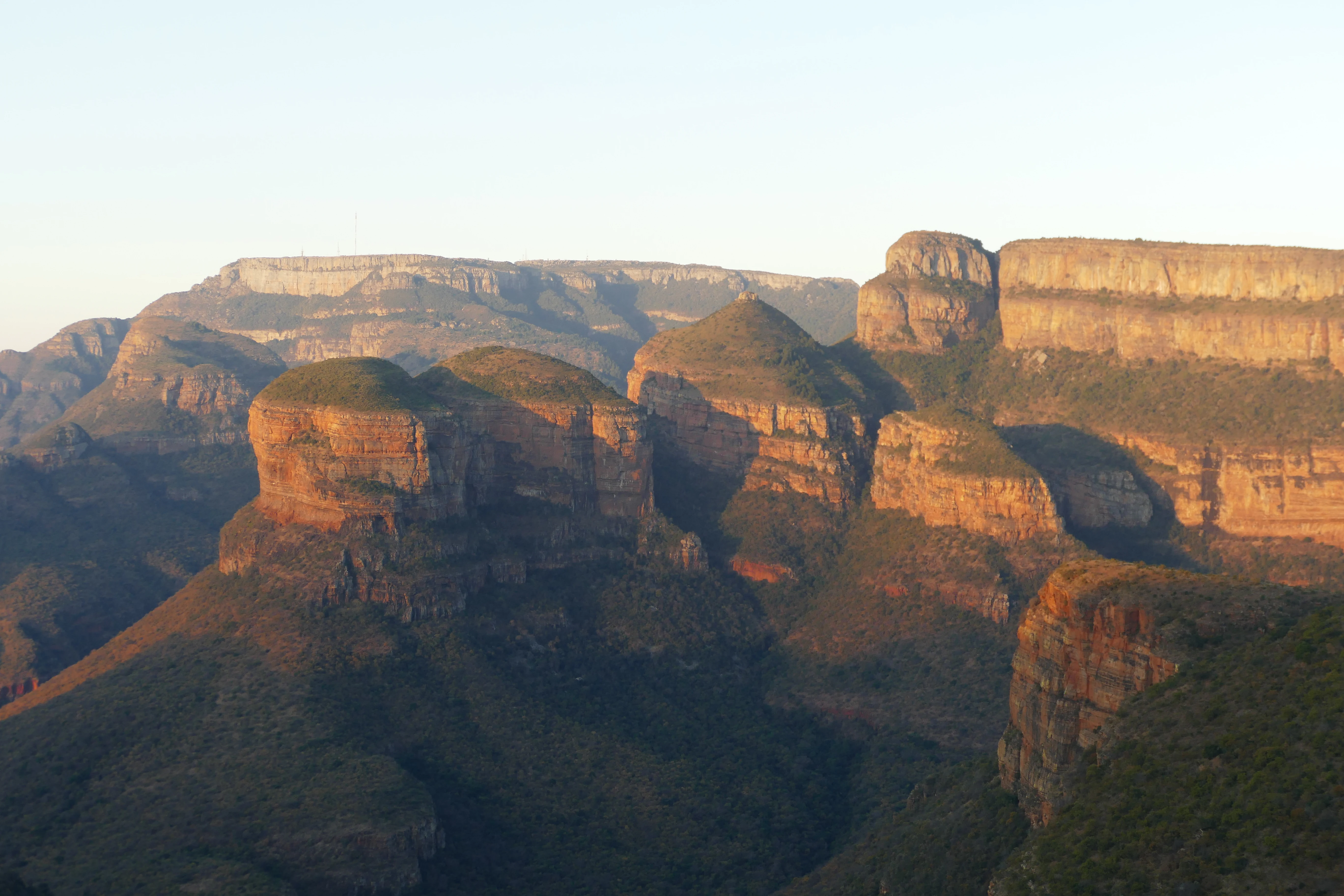 A wide view of the Three Rondavels, three prominent hut-shaped mountain peaks, illuminated by golden hour light against a deep canyon landscape.
