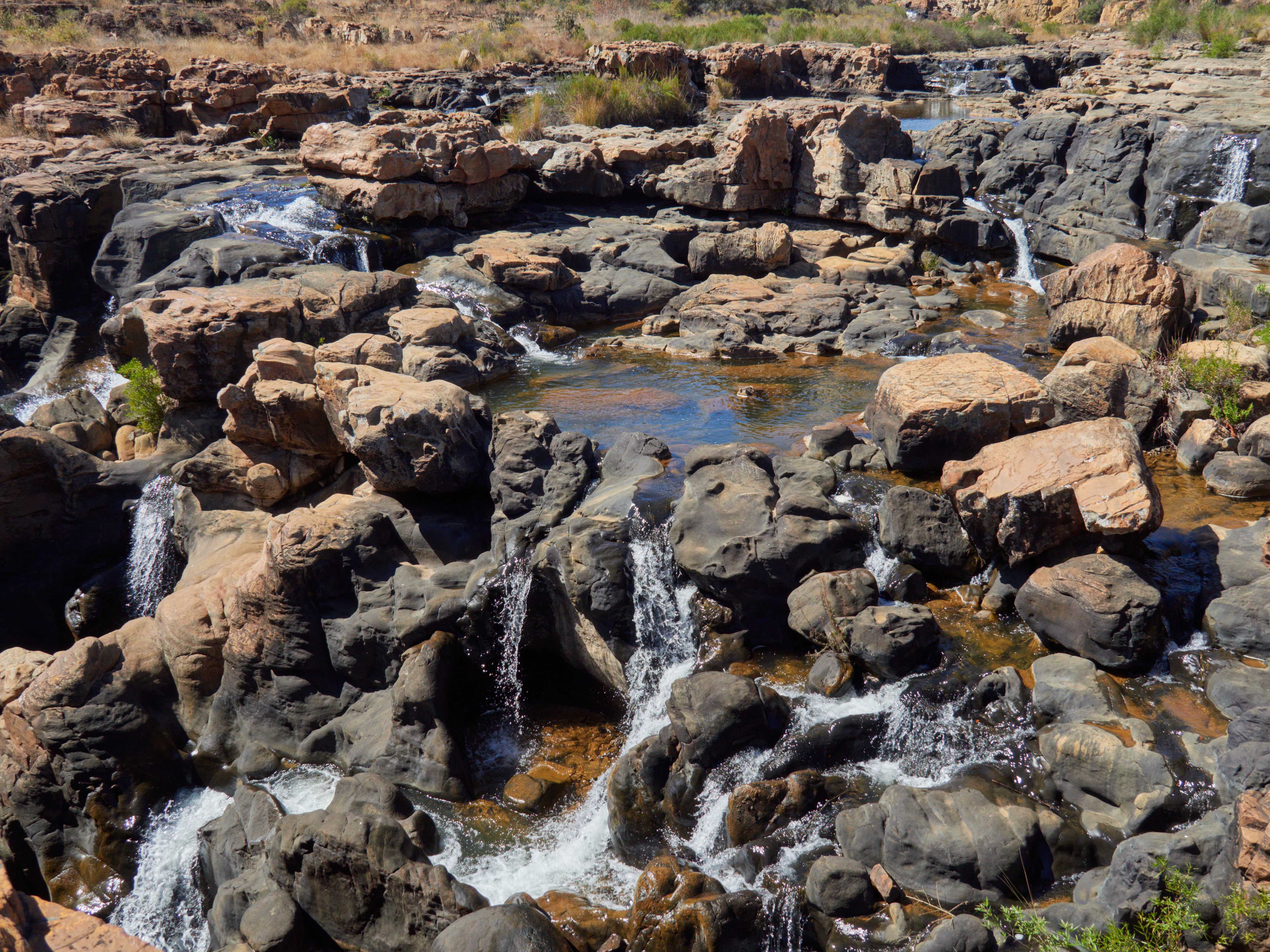 A high-angle view of rugged, dark, eroded rock formations with small waterfalls and streams of water flowing into pools at Bourke's Luck Potholes.