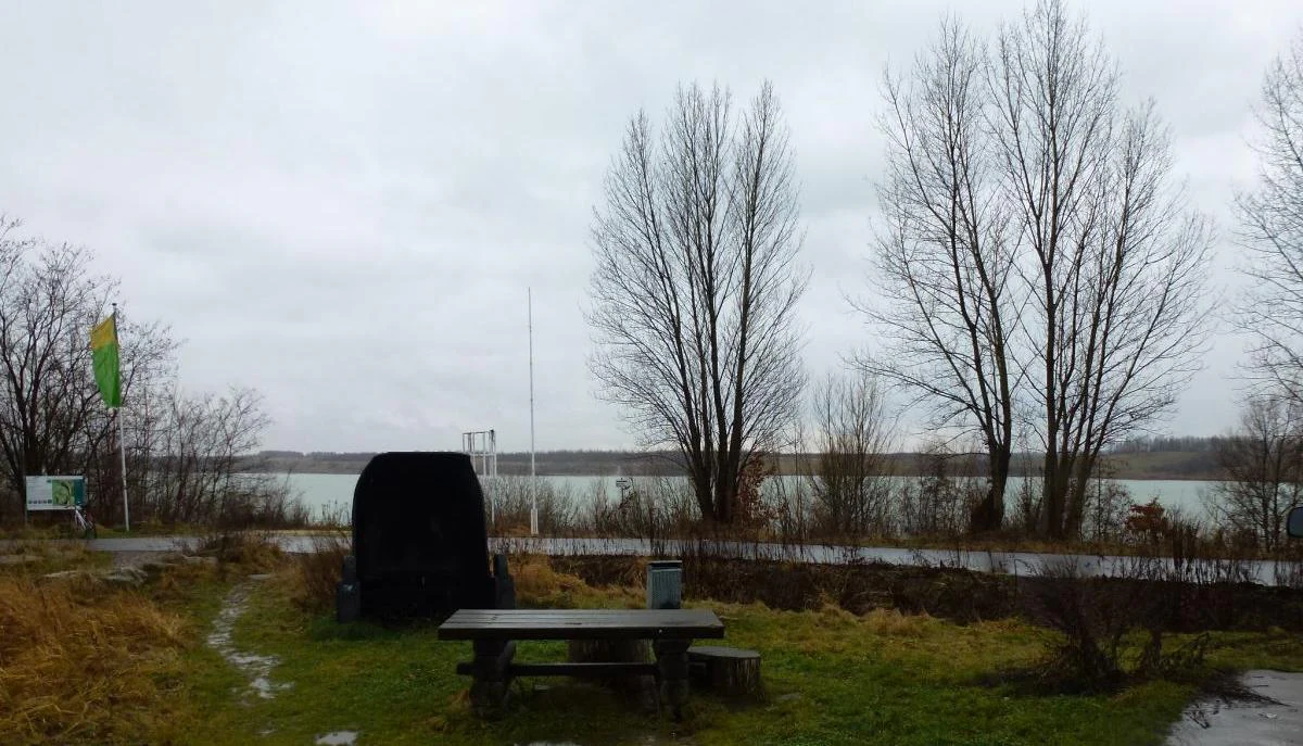 A outdoor bench sits on a grassy area in front of a wide lake under an overcast sky.