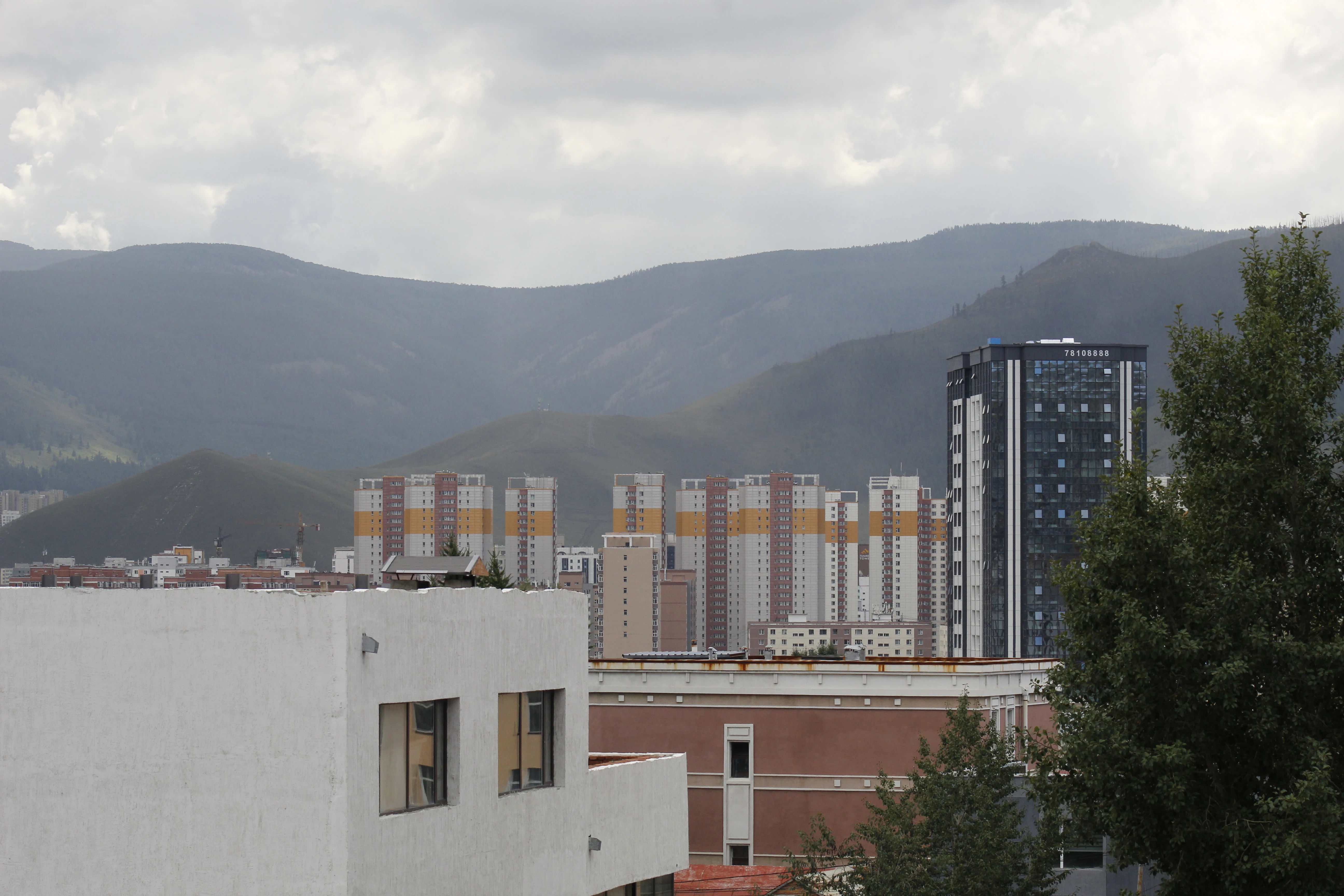A view of the rolling slopes of Bogd Khan Uul rising behind the high rise apartment buildings and urban structures of Ulaanbaatar under a cloudy sky.