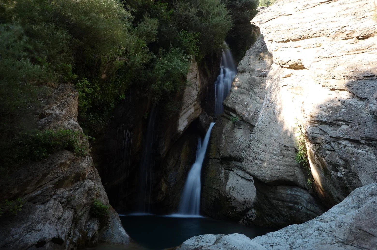A multi-tiered waterfall flows down a rocky cliffside into a pool, framed by trees on one side and light-colored rock walls on the other.