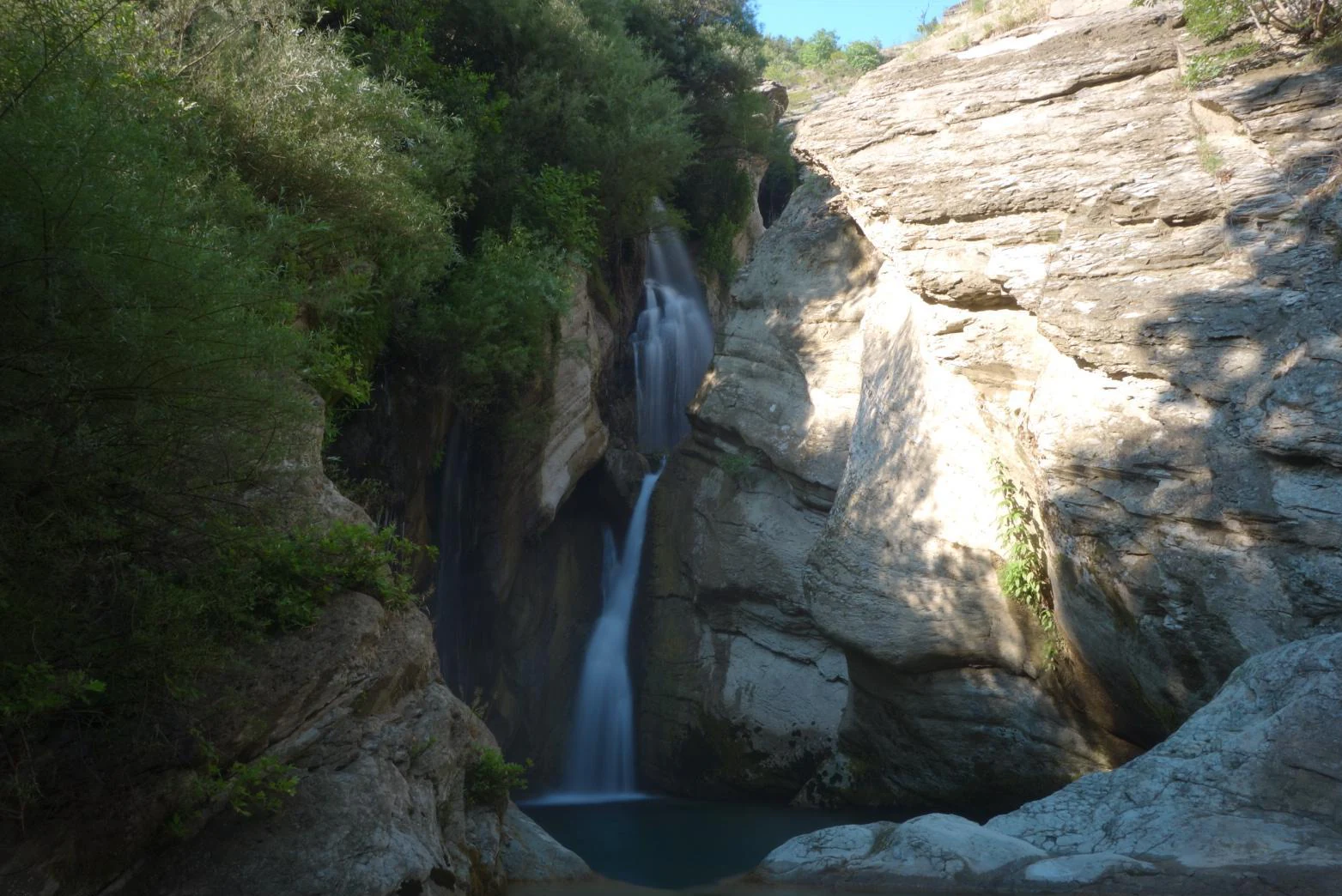 A waterfall cascades down a rocky canyon wall surrounded by lush greenery and shaded cliffs.