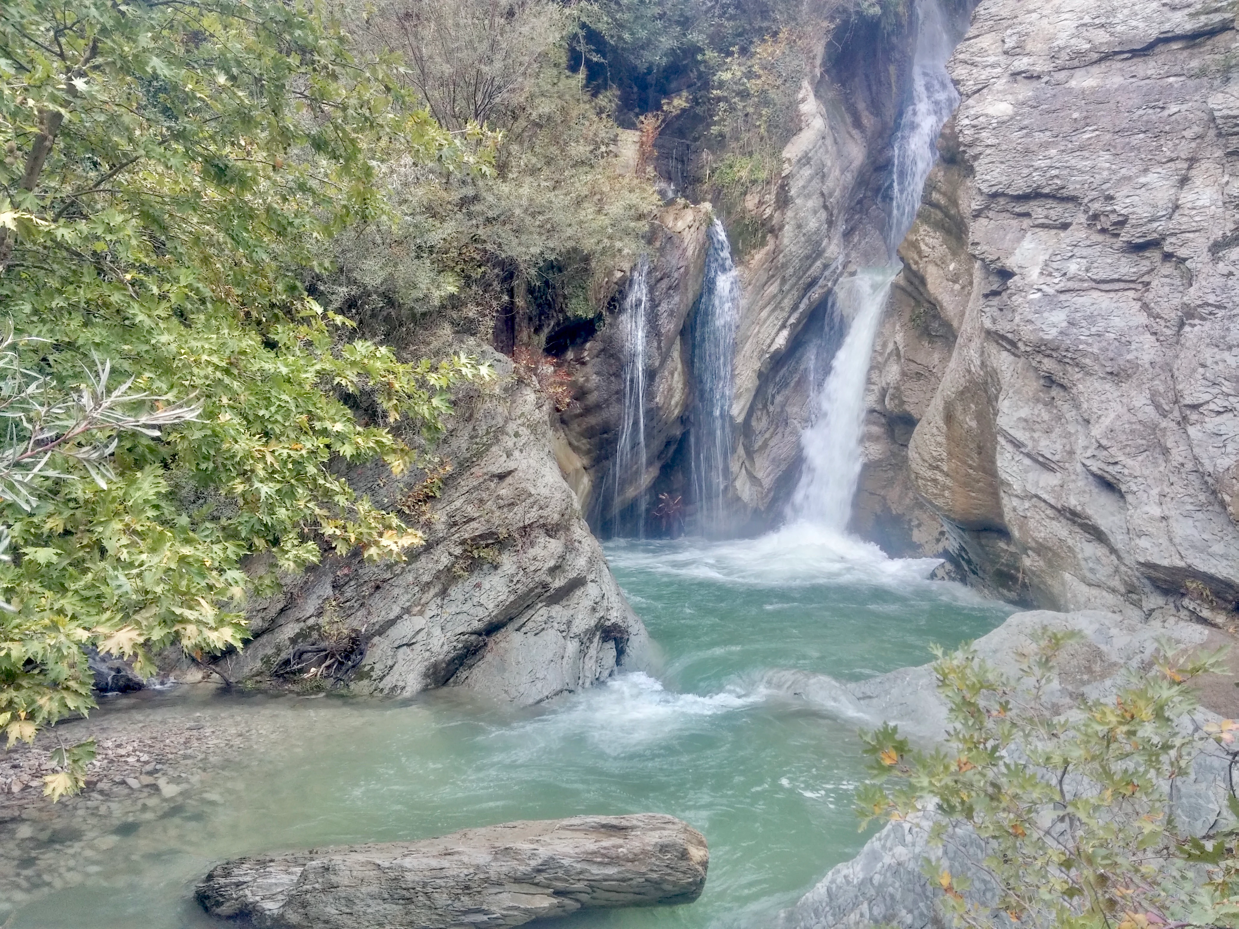 A multi-tiered waterfall cascading down rocky cliffs into a turquoise stream, surrounded by lush green trees and foliage.