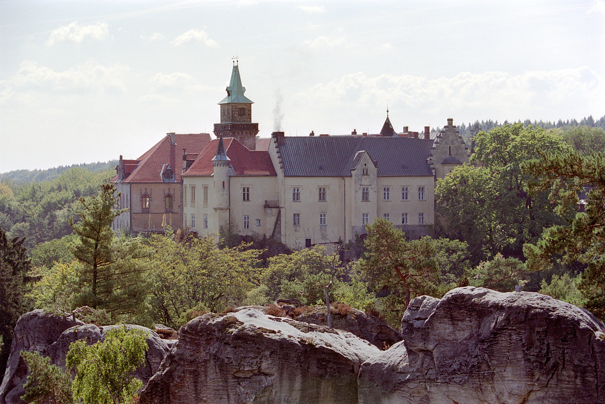 A large castle with red and dark gray roofs, surrounded by green trees and sandstone rock formations, under a partly cloudy sky