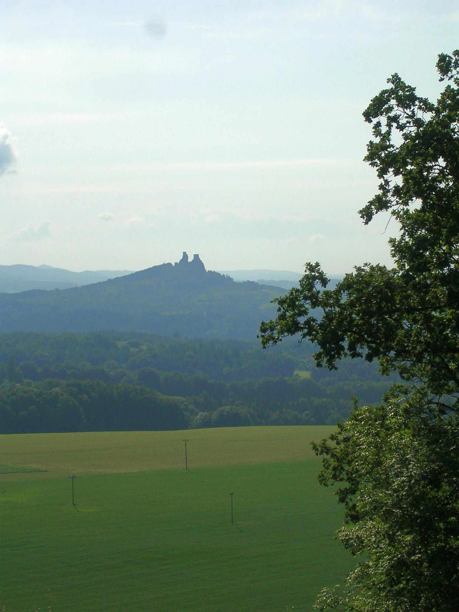 Landscape view showing Trosky Castle ruins in the distance with green fields and forested hills in the foreground, viewed from Vyskře, Czech Republic.