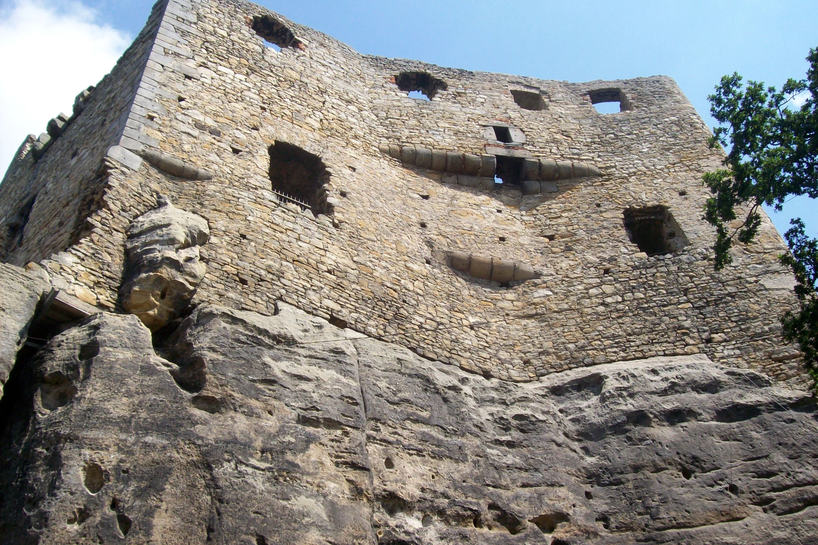 Stone castle ruins with multiple openings carved into a rocky cliff face under a blue sky