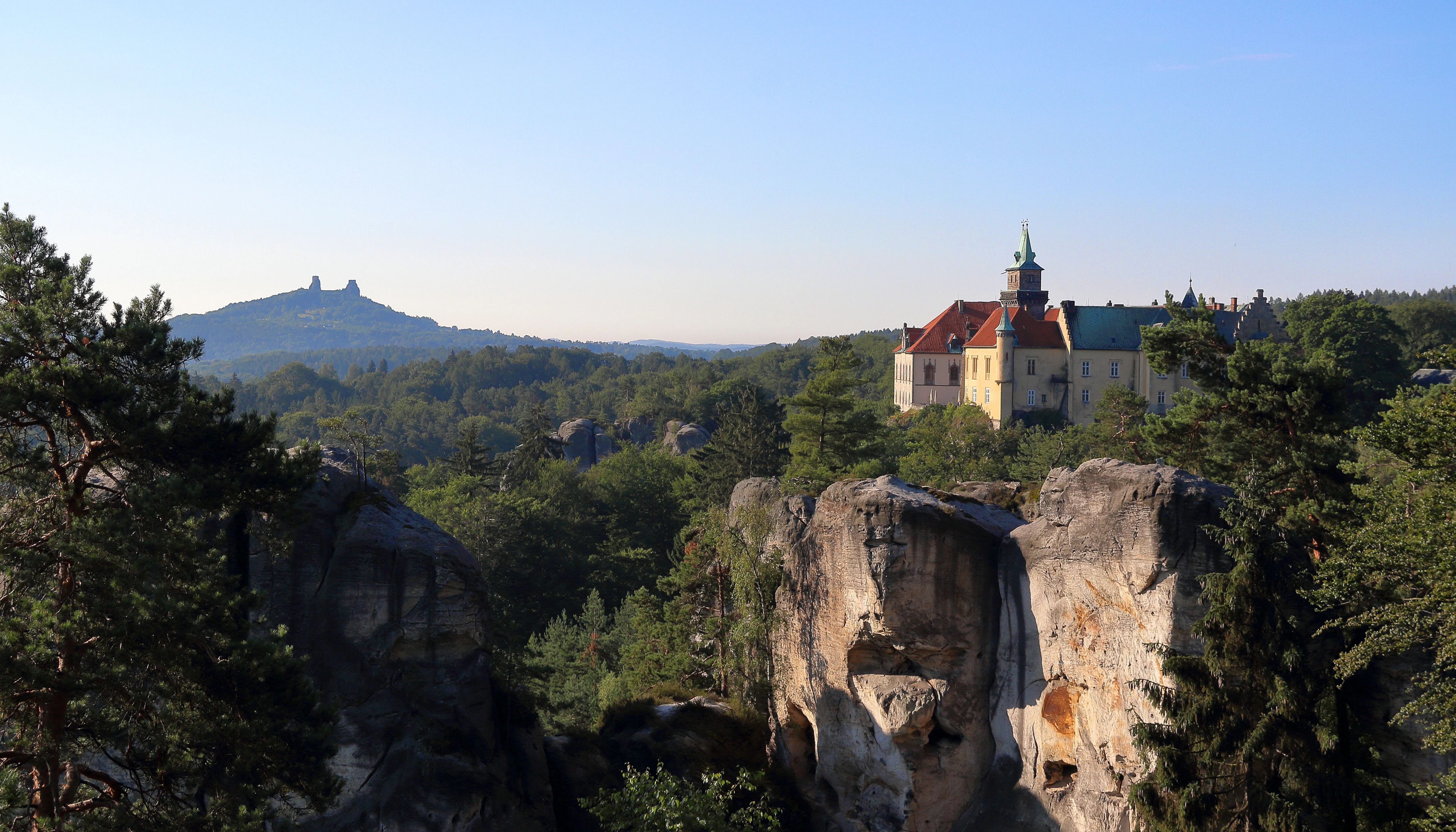Panoramic view of sandstone rock formations with Castle Hruba Skala on a rock outcrop, surrounded by forested hills under clear blue sky