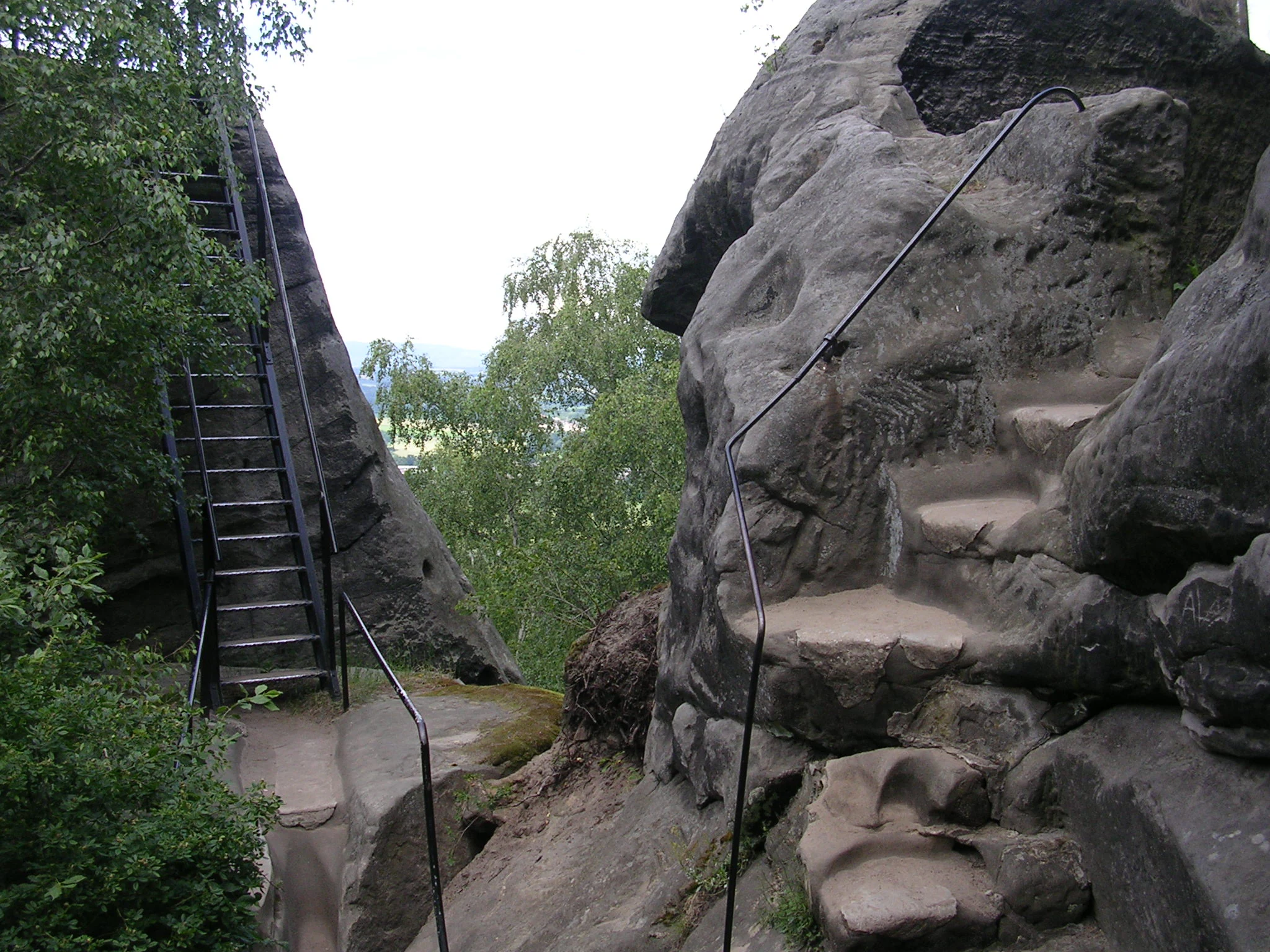 Metal staircase and railings on a rocky cliff face with green vegetation and distant landscape view