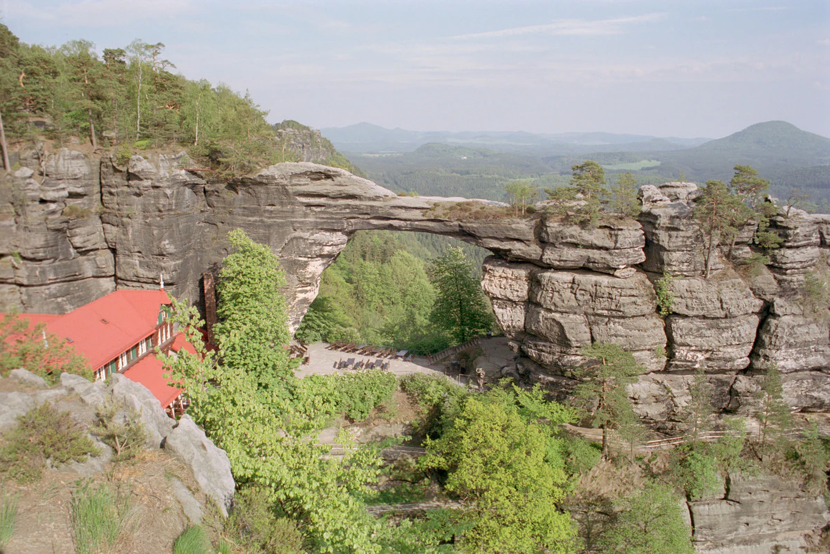 Natural sandstone arch spanning a valley with rocky cliffs, green trees, and a red-roofed building on the left side.