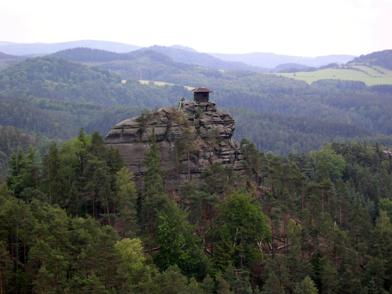 Rock formation with small structure on top, surrounded by dense forest and rolling hills