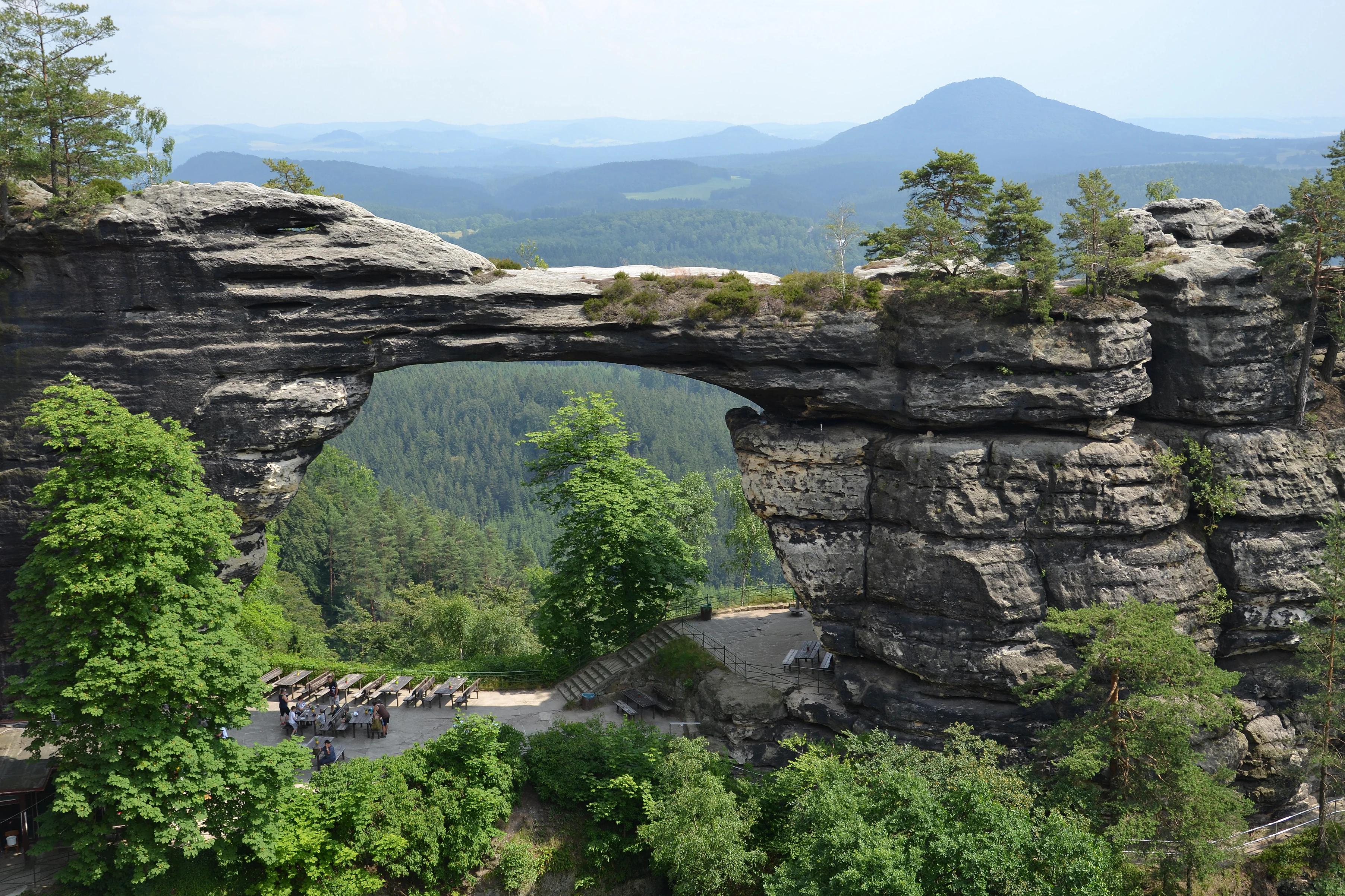A large natural sandstone arch spanning a valley with green trees and distant mountains
