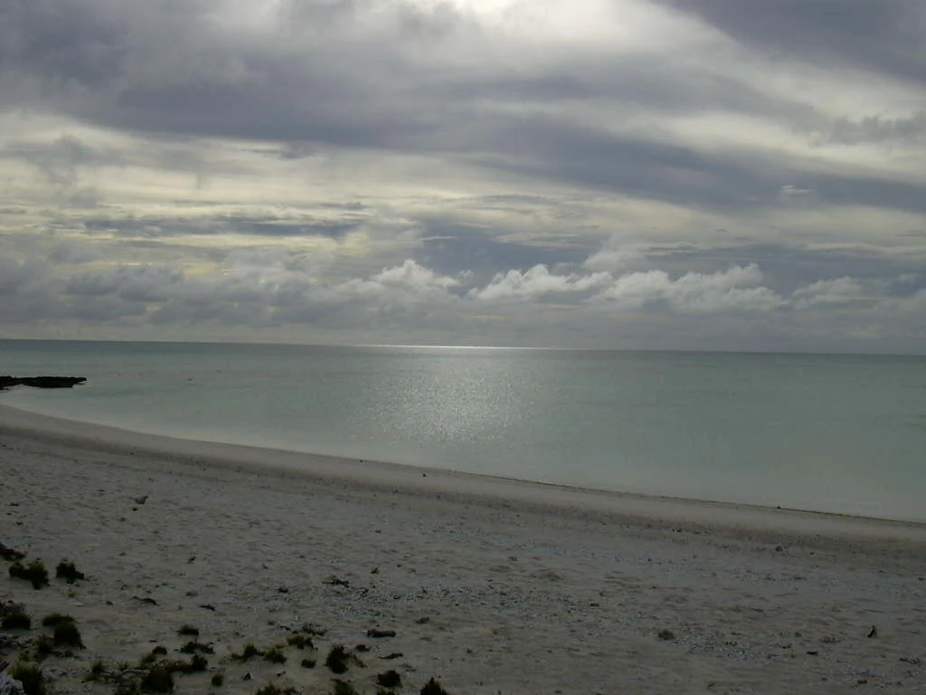 Una vista gran angular de una playa de arena blanca que se encuentra con las aguas tranquilas y poco profundas de una laguna bajo un cielo nublado.