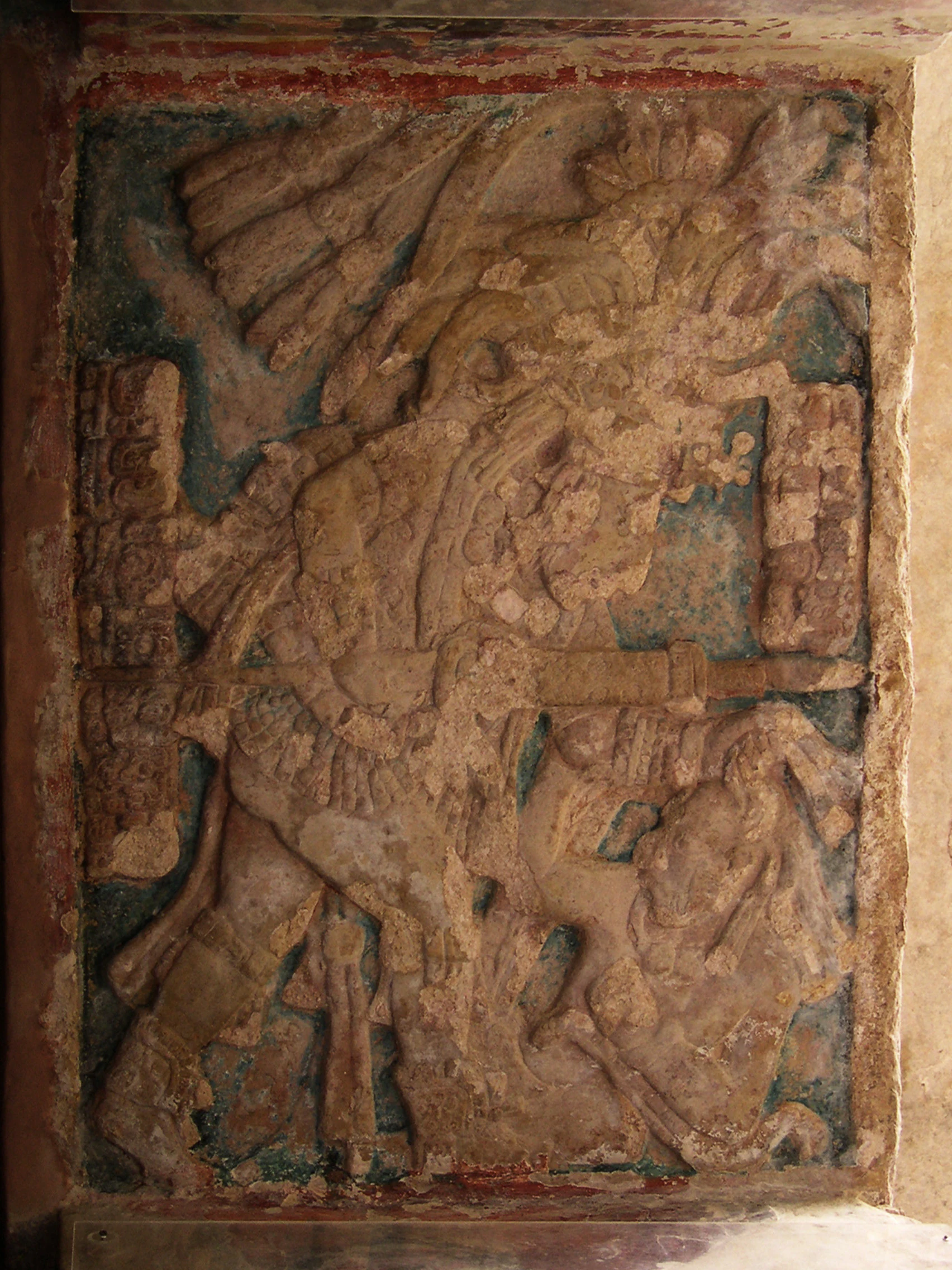An ancient stone lintel relief carving featuring a Maya figure in elaborate headdress and clothing, with surrounding glyphic script and decorative elements.