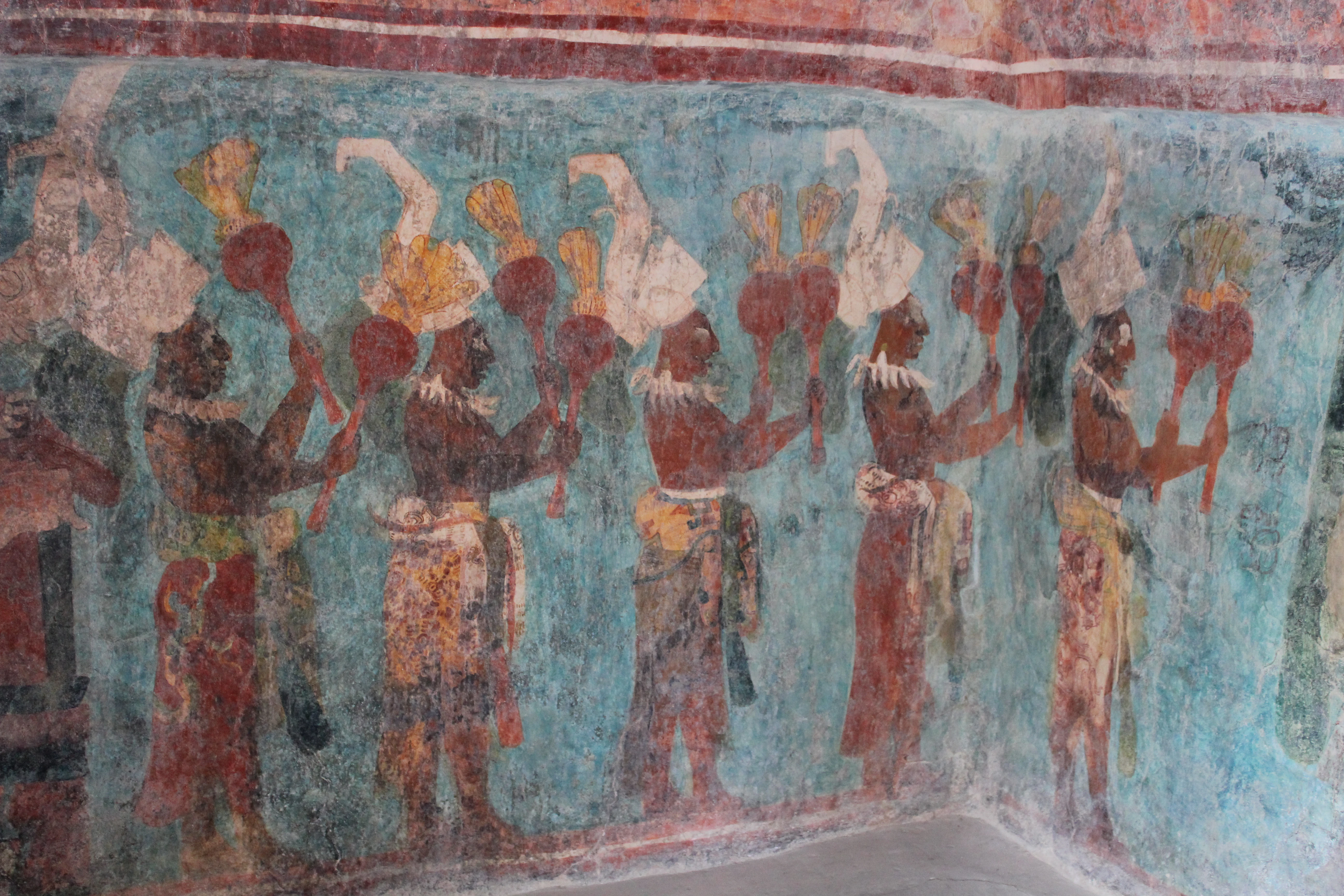 A section of an ancient Maya wall mural depicting a procession of figures holding instruments against a blue background.