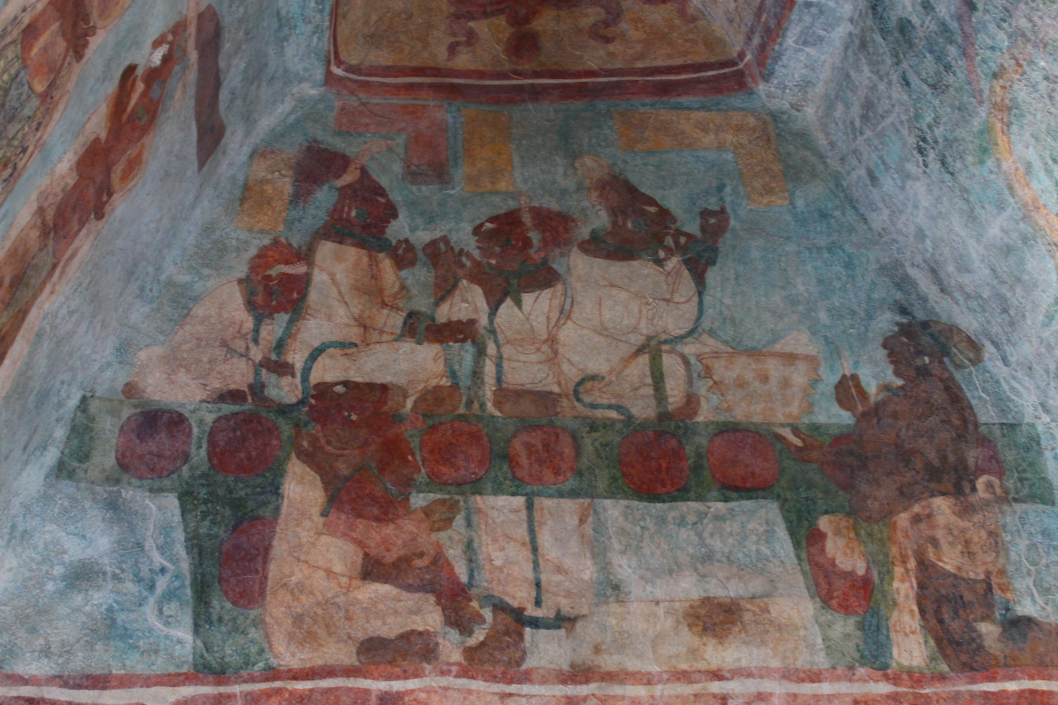 A faded ancient Mayan mural on a stone wall depicting several figures, likely dignitaries, seated at a table or dais.
