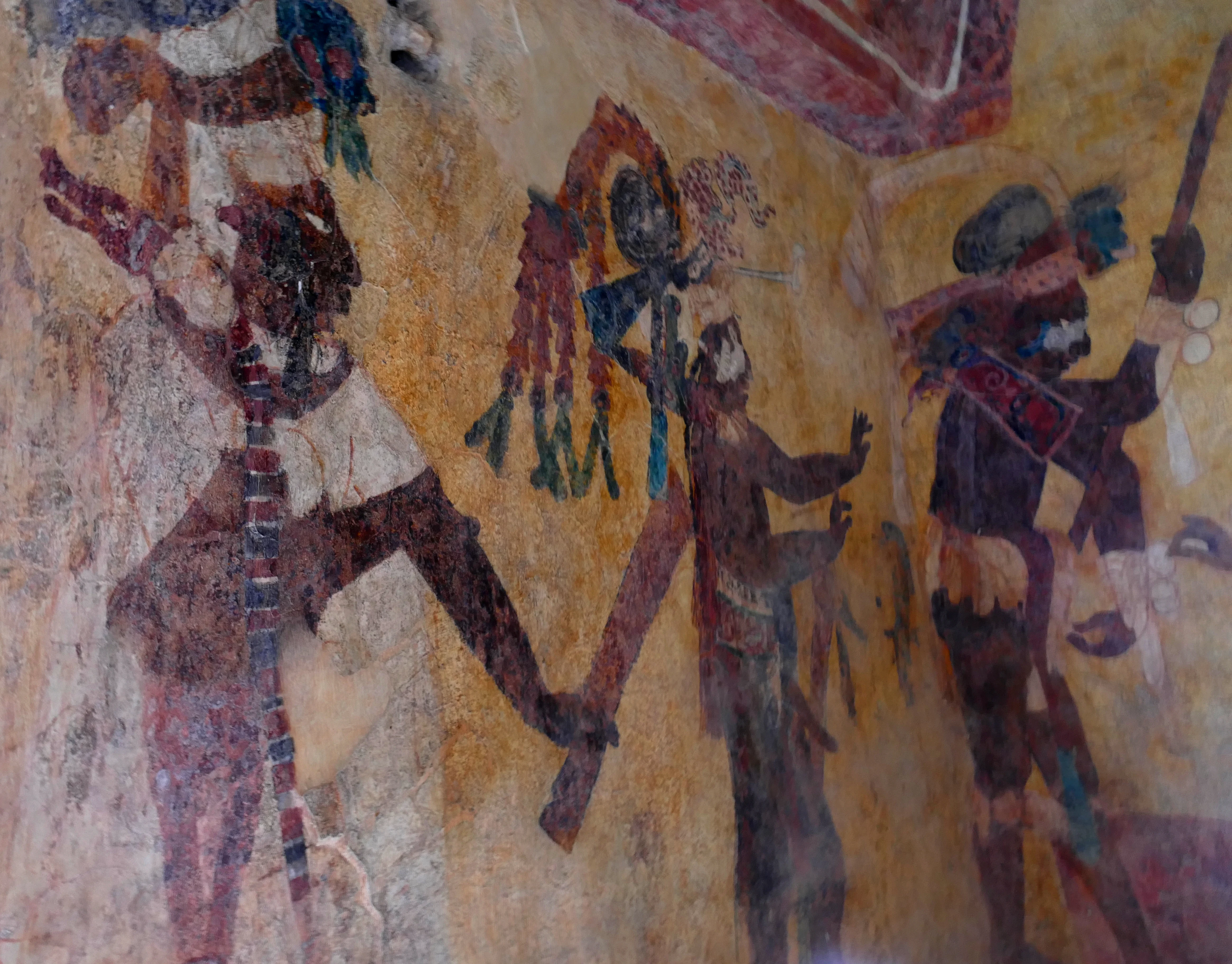 A close up view of faded ancient Mayan wall paintings depicting figures in ceremonial attire and headdresses in warm ochre and earth tones.