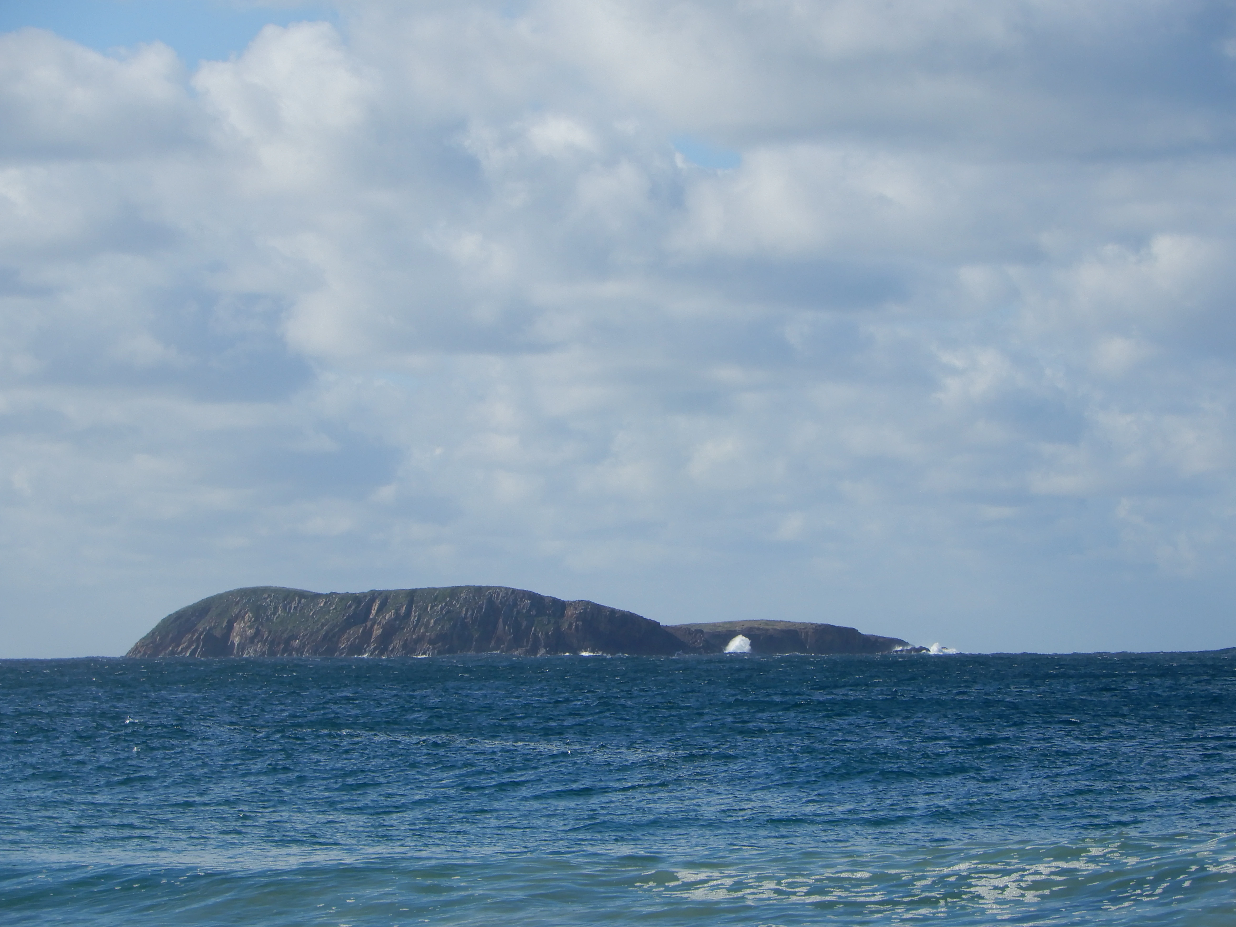 A low, rocky island sits on the horizon in the middle of a calm ocean under a partly cloudy sky.
