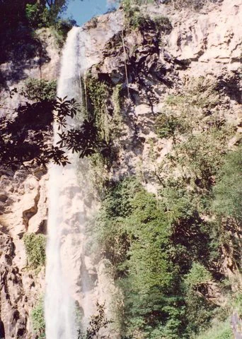 A tall, narrow waterfall plunges down a steep, rocky, tree-covered cliff face.
