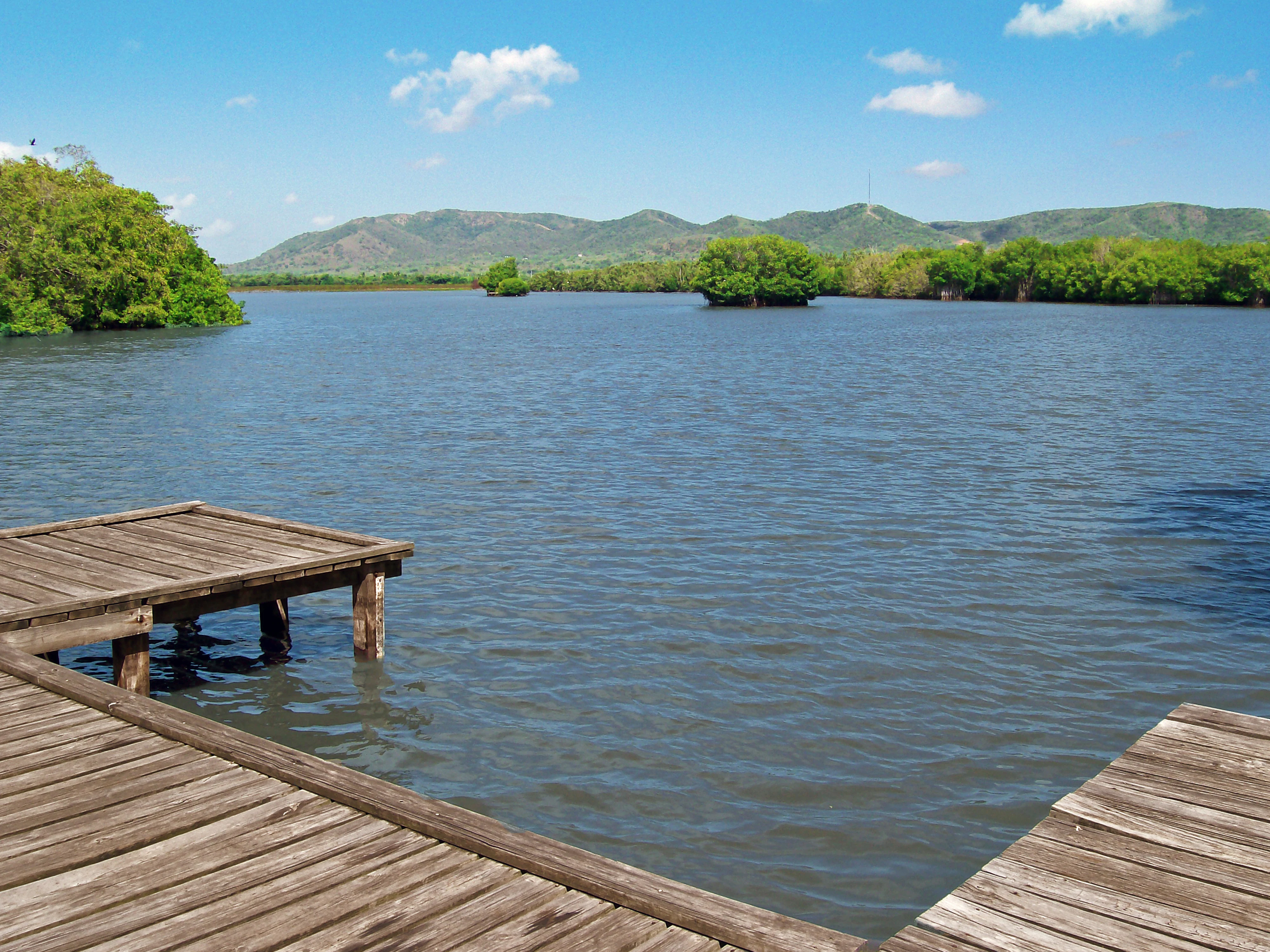 A wooden boardwalk platform extends into the calm, dark waters of a lagoon, with mangrove-lined shores and rolling green hills in the background under a blue, partly cloudy sky.
