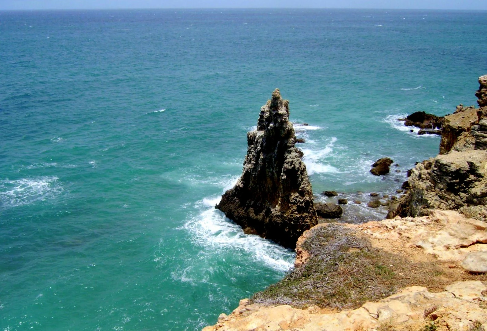 A jagged, vertical limestone rock pillar rises from turquoise ocean waters near the base of rocky coastal cliffs.