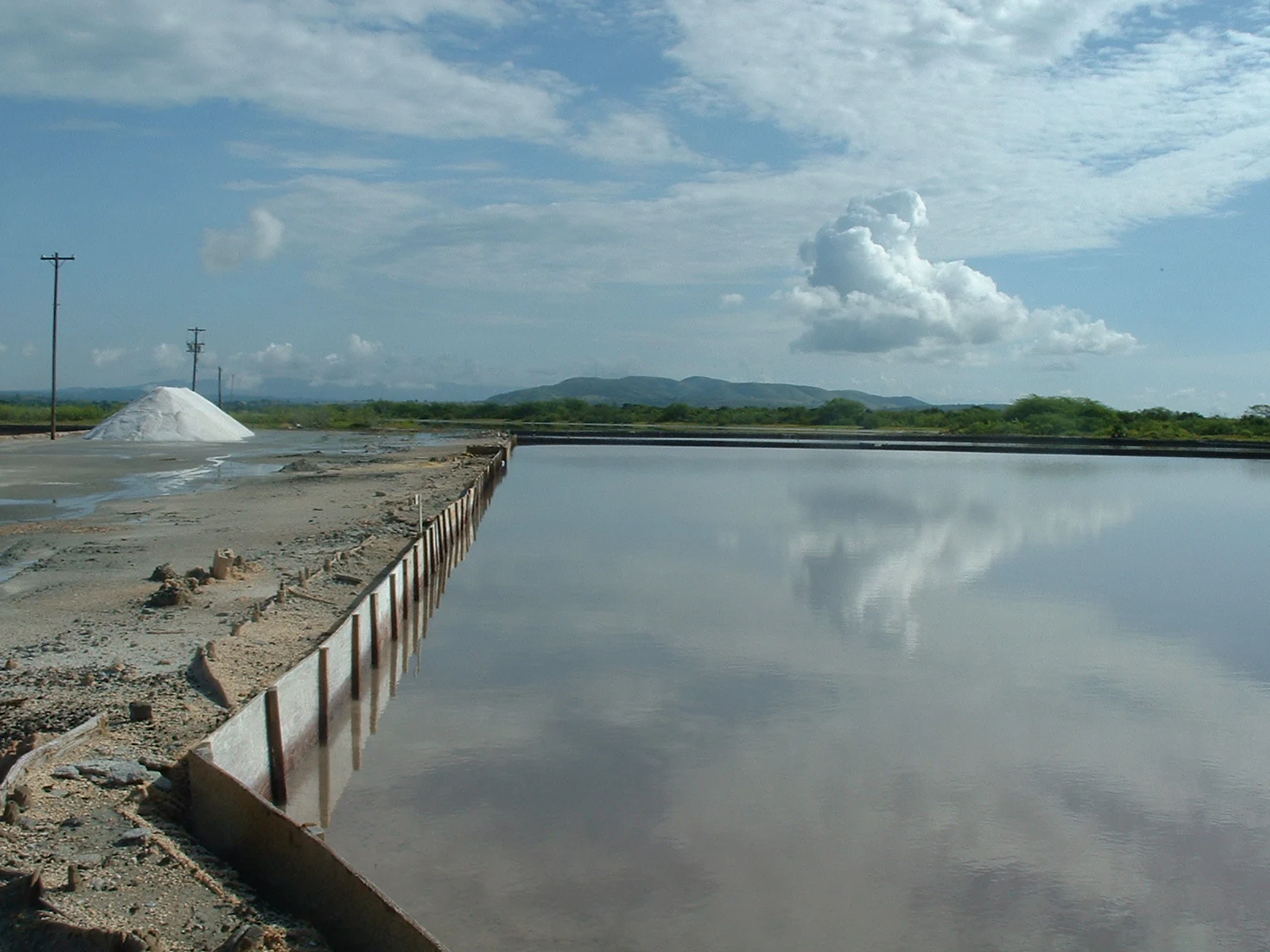 Wide shot of the Salinas de Cabo Rojo salt flats with a large pile of harvested salt on the left and a reflective pool of water mirroring the blue sky and clouds.