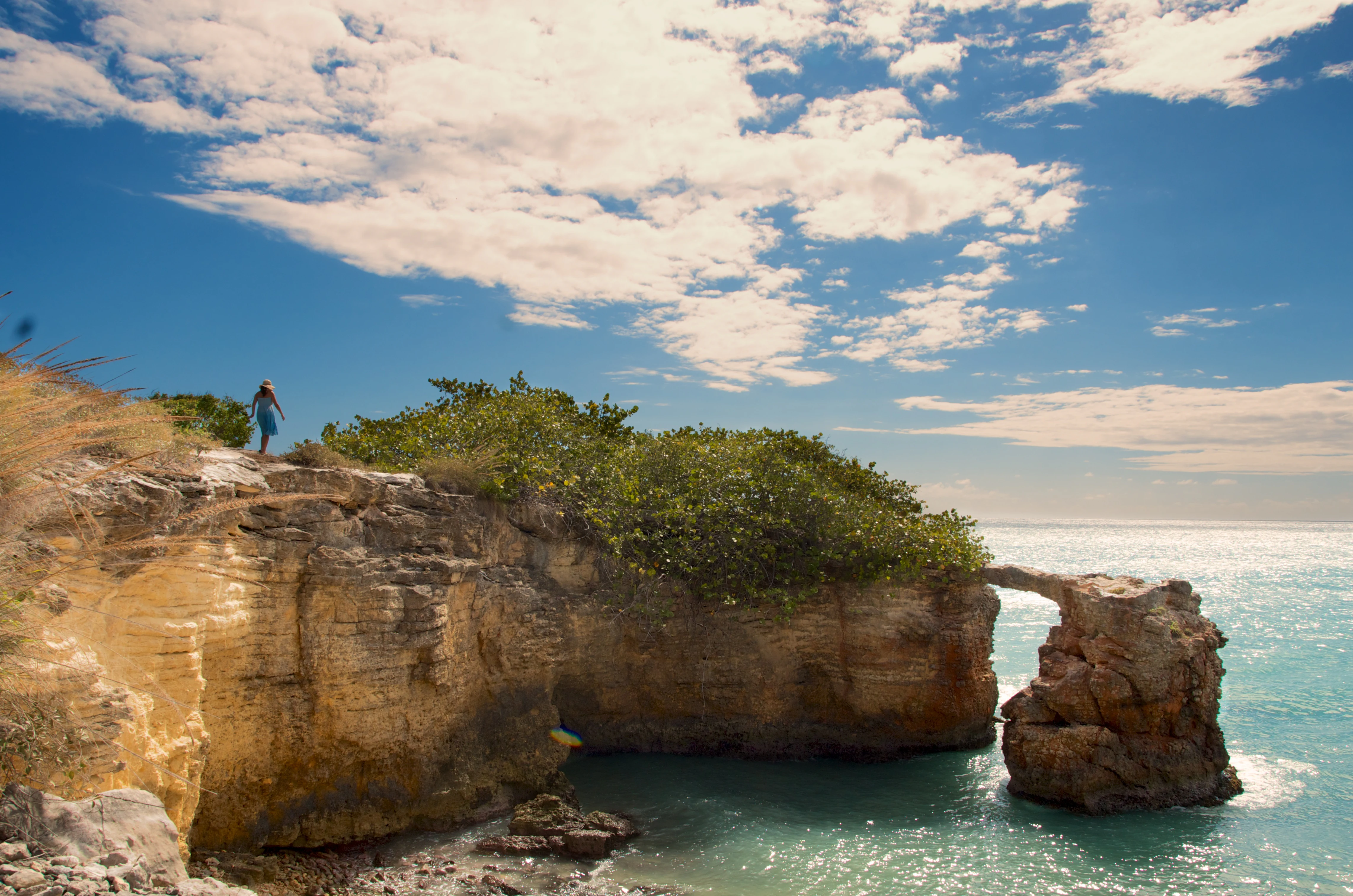 A coastal landscape view showing steep golden limestone cliffs with low vegetation overlooking turquoise ocean water with a natural rock arch formation.