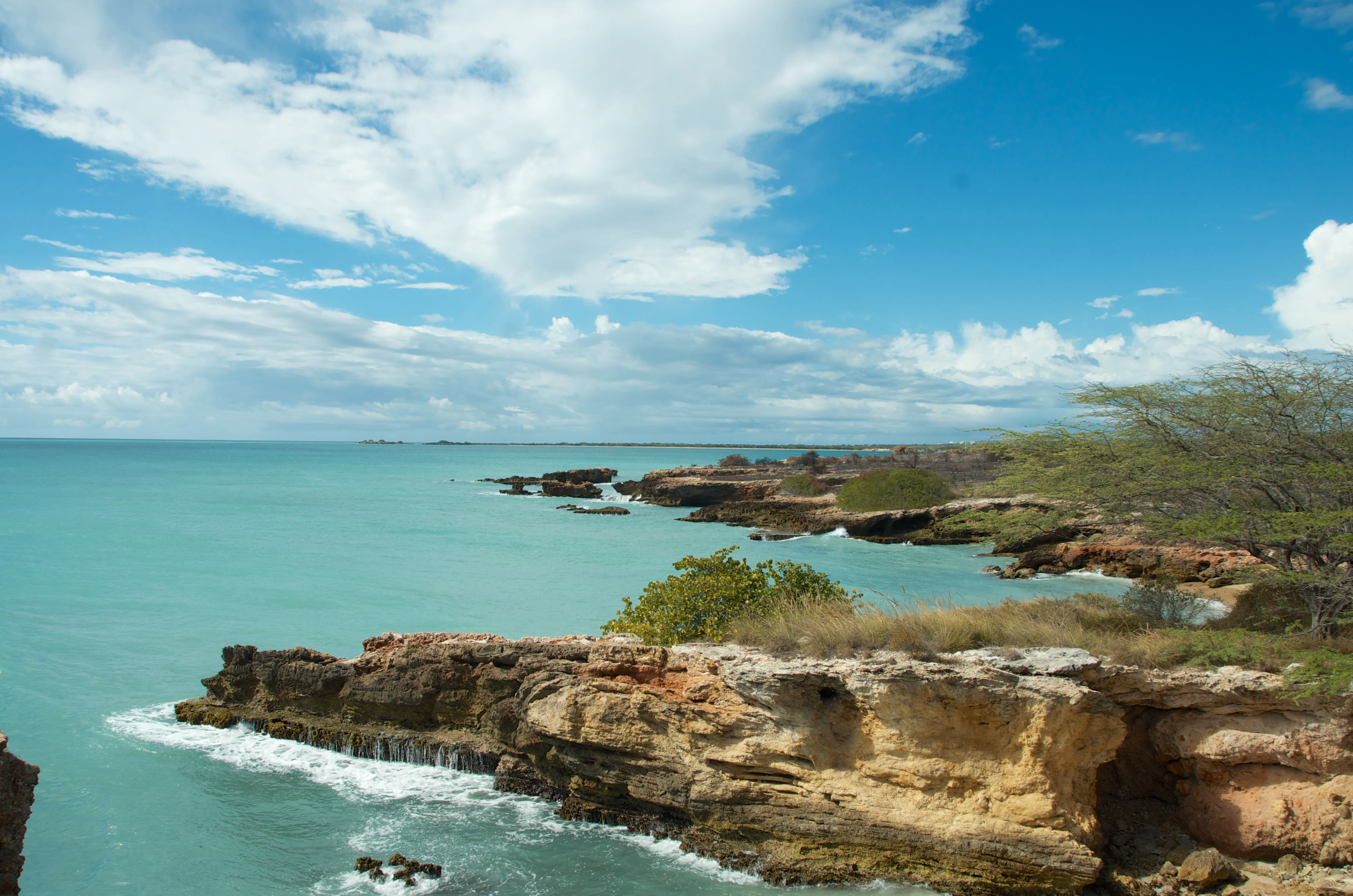 A wide angle view of a turquoise Caribbean sea meeting rugged, tan limestone cliffs under a bright blue sky with scattered white clouds.