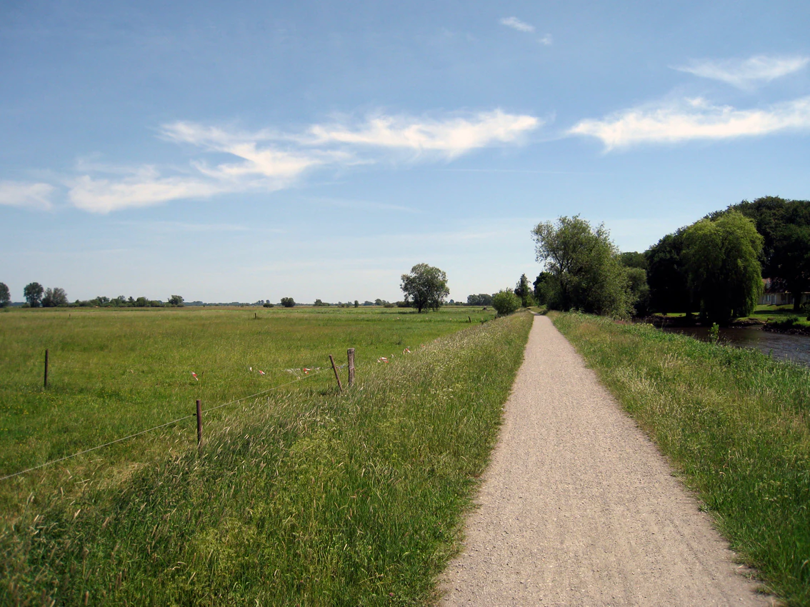 A wide, gravel walking path leads into the distance, with green meadows on the left and trees along a river bank on the right under a blue, partly cloudy sky.