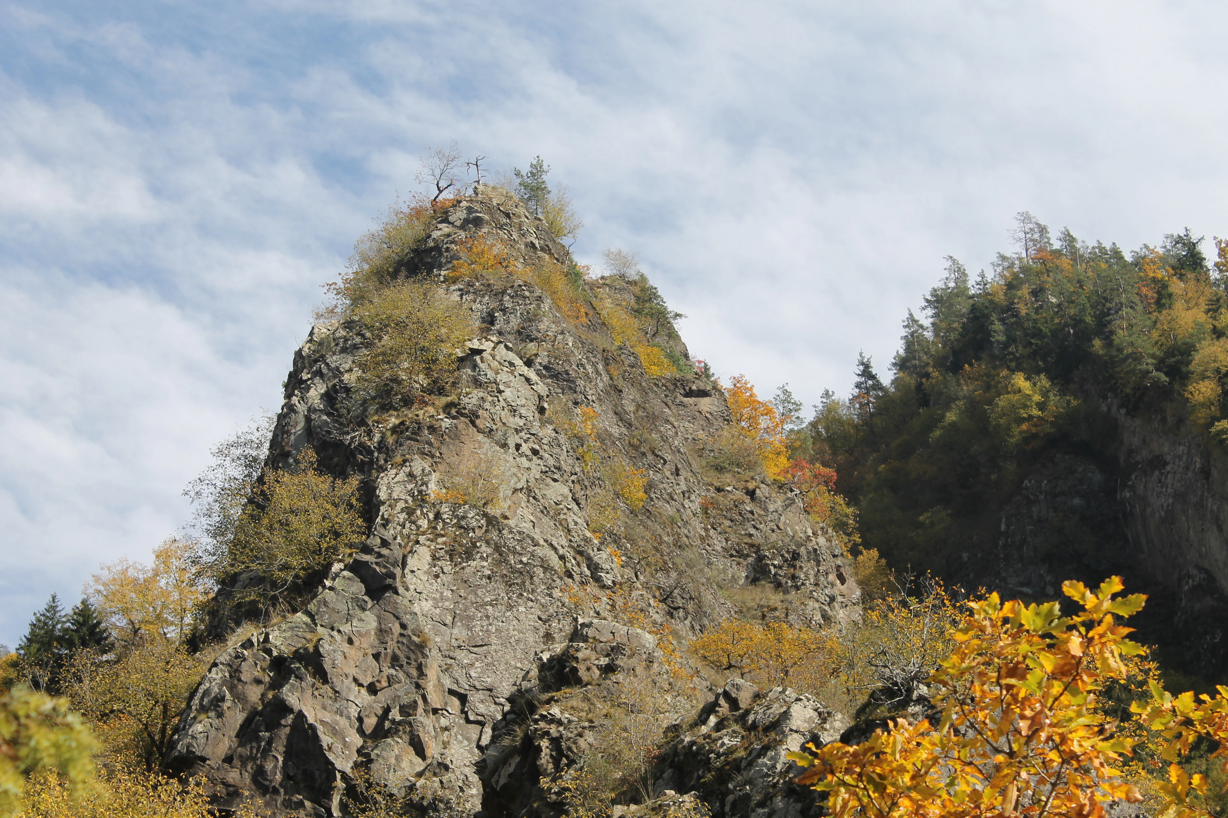 Rocky mountain peak with sparse vegetation and autumn-colored trees under a partly cloudy sky