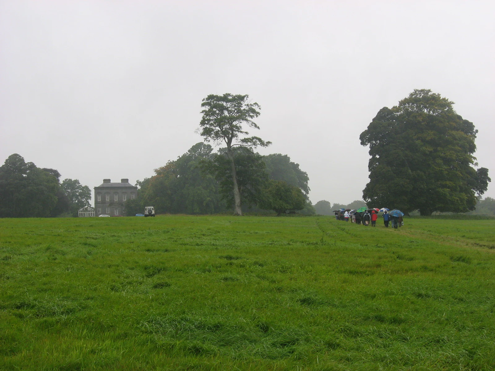 A green, grassy field under an overcast sky with large trees and a distant stone house. A small group of people with umbrellas is walking on the right side of the frame.