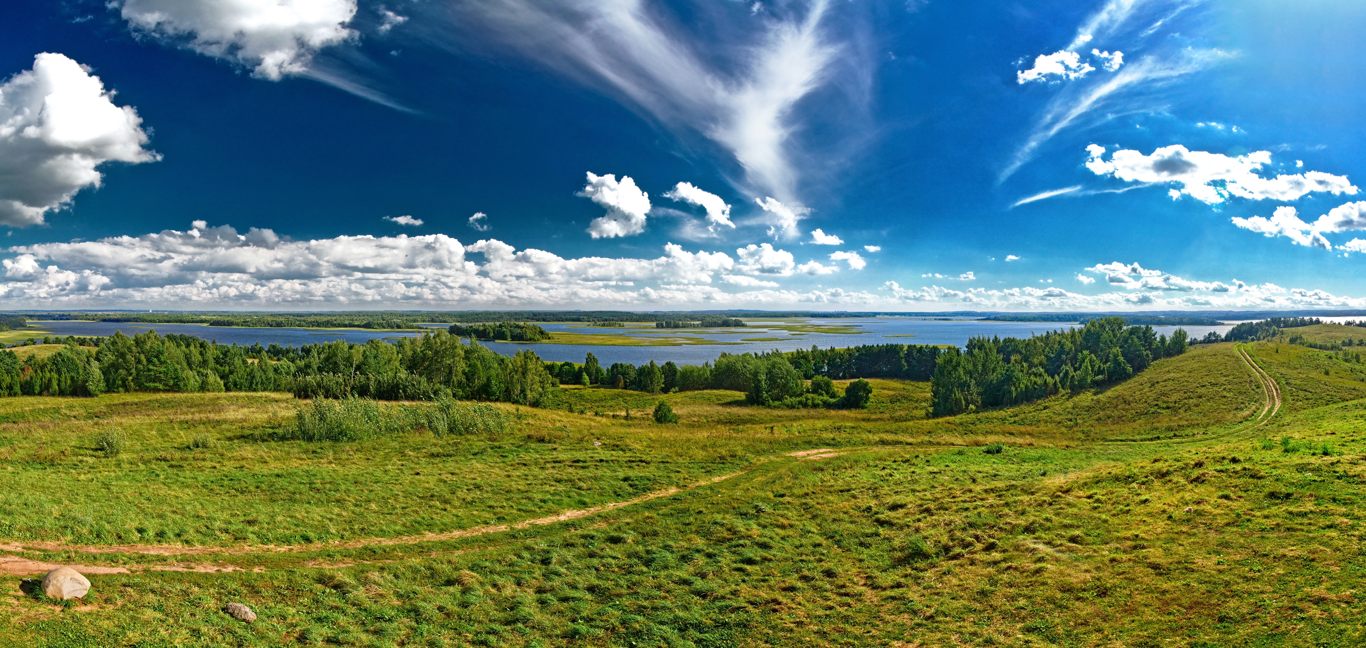 Panoramic landscape showing Strusta Lake, green meadows, scattered trees, and a blue sky with clouds
