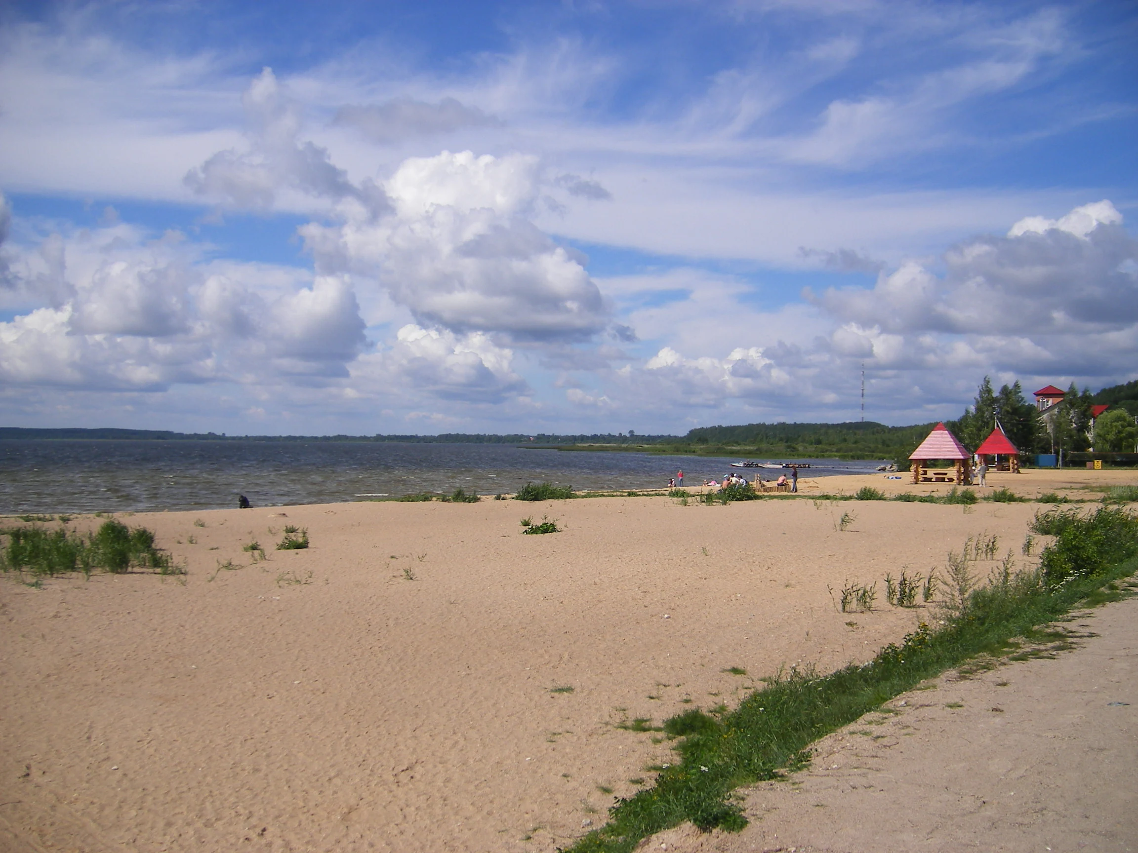 Sandy beach with grass patches and a dirt path leading to a lake shore. Small red-roofed structures are visible on the right side. The lake extends into the distance with a partly cloudy sky above.