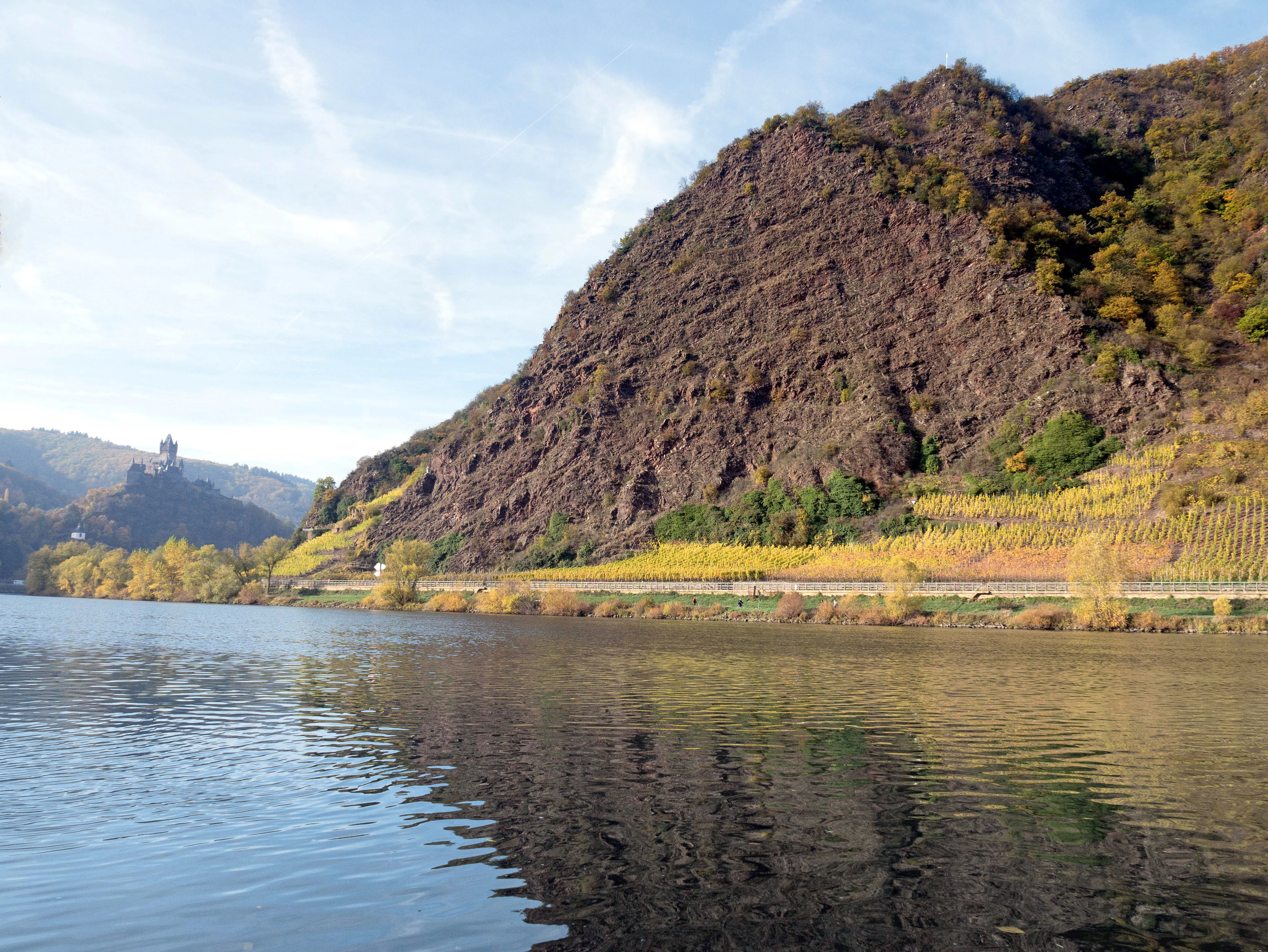 A steep, dark, rocky cliff face slopes down toward the Moselle River, which reflects the sky. Yellow-leaved vineyards line the base of the rock, and a castle is visible on a distant hillside.