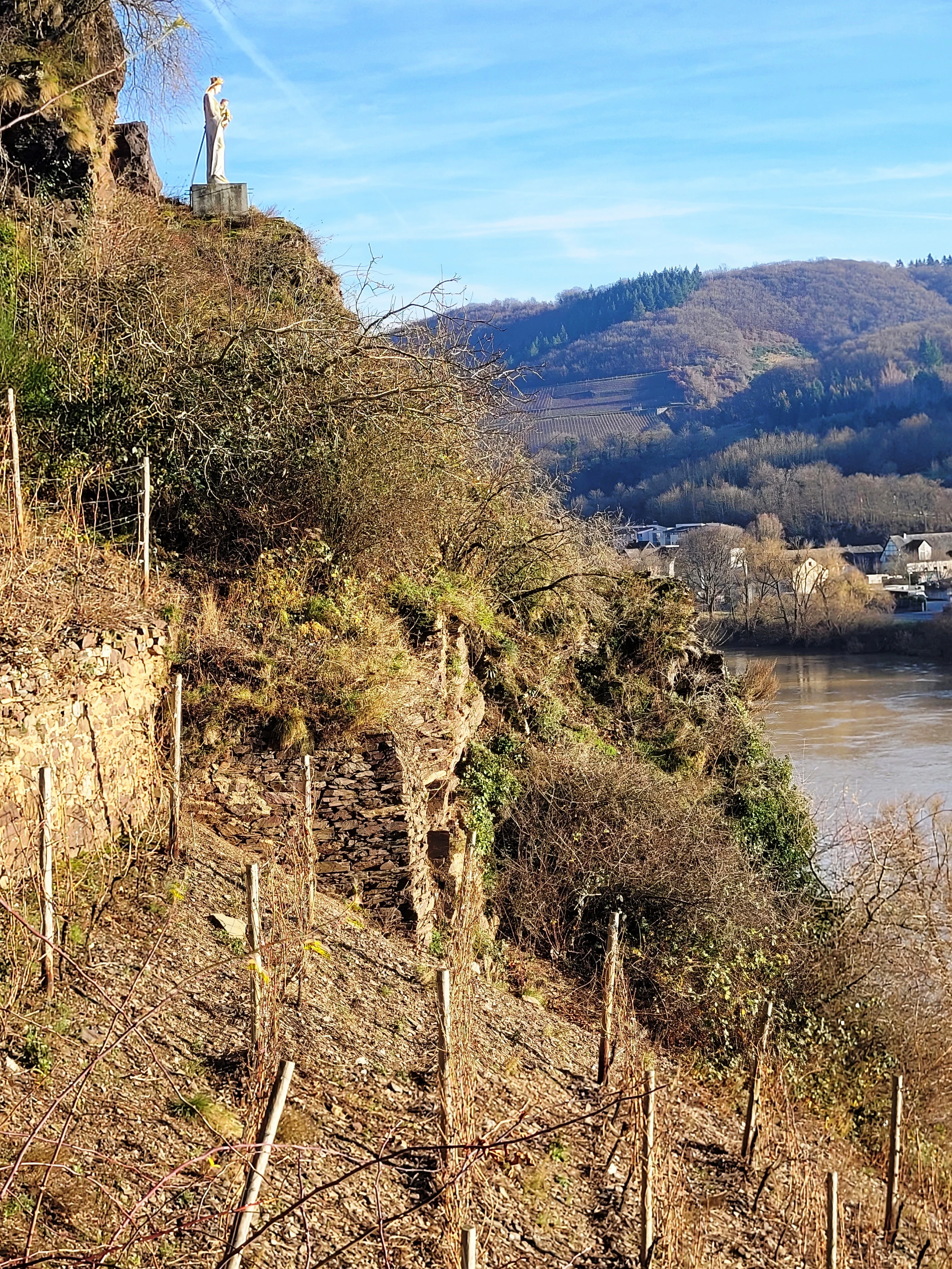 A white statue of a figure stands on a rocky outcrop above a steep slope overlooking a river valley.
