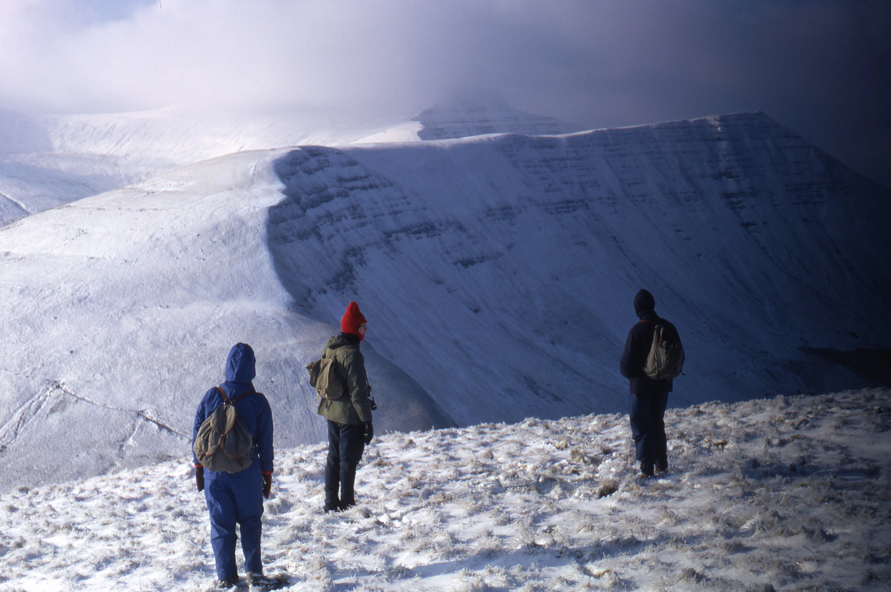 Three people standing on a snowy mountain slope with rocky peaks in the distance.