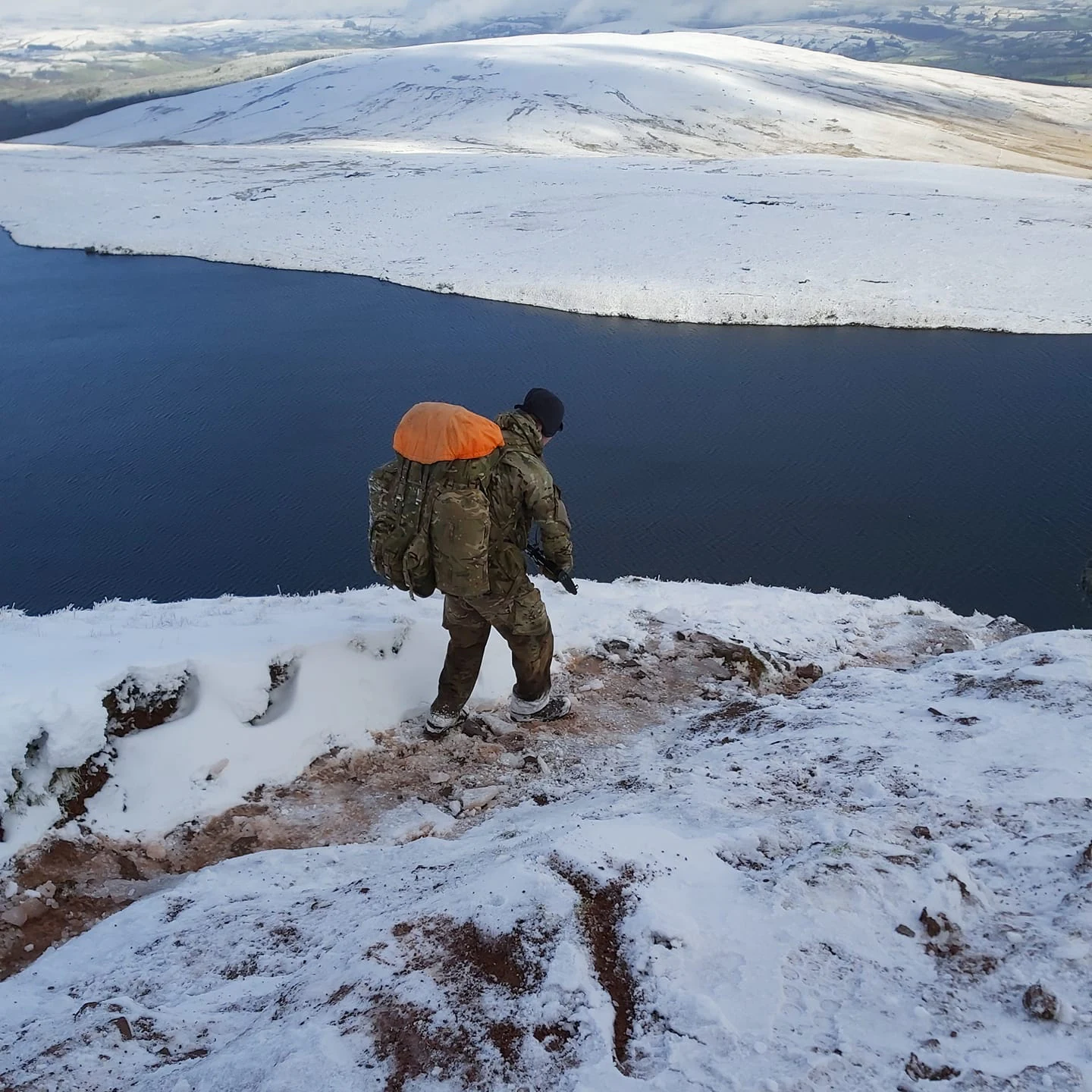 Military personnel in camouflage uniform with orange backpack standing on snow-covered hillside above a lake