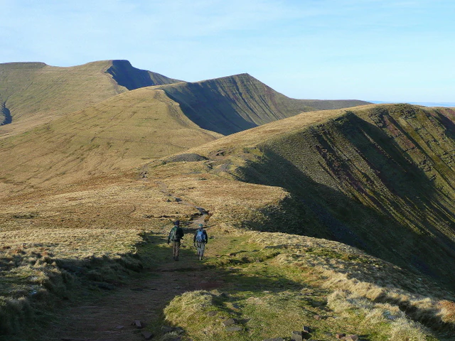 Two hikers walking on a trail across a grassy mountain ridge with rolling hills and peaks under a clear sky.