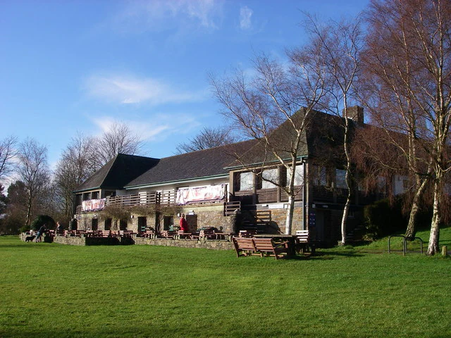 Stone and timber visitor centre building with sloped roof, outdoor benches, and large grassy area under a clear blue sky